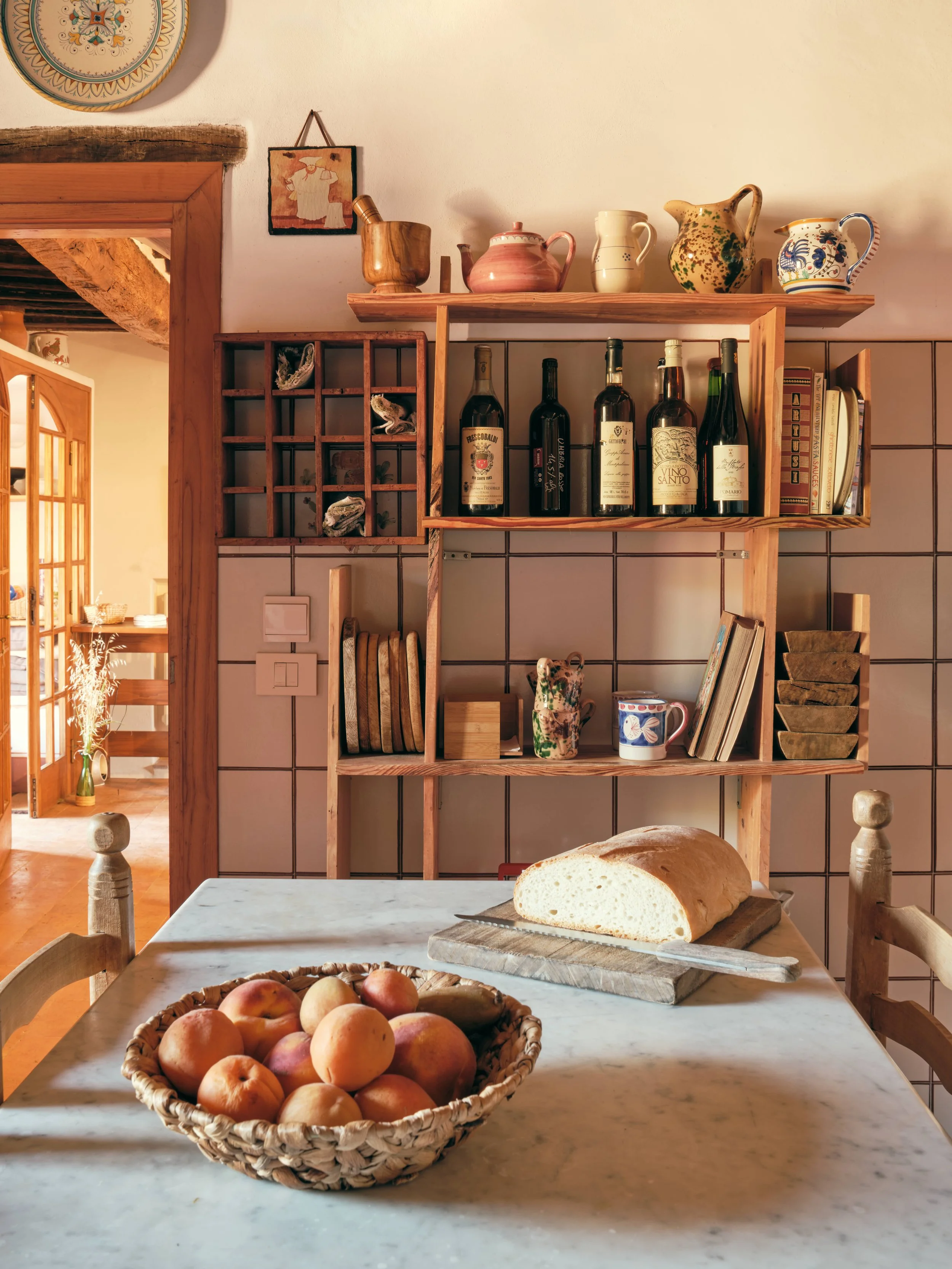 Bowl of peaches and nectarines on a marble kitchen table, with a loaf of bread and a bread knife in the foreground, and kitchen shelves with bottles, cups, and decorative items in the background.