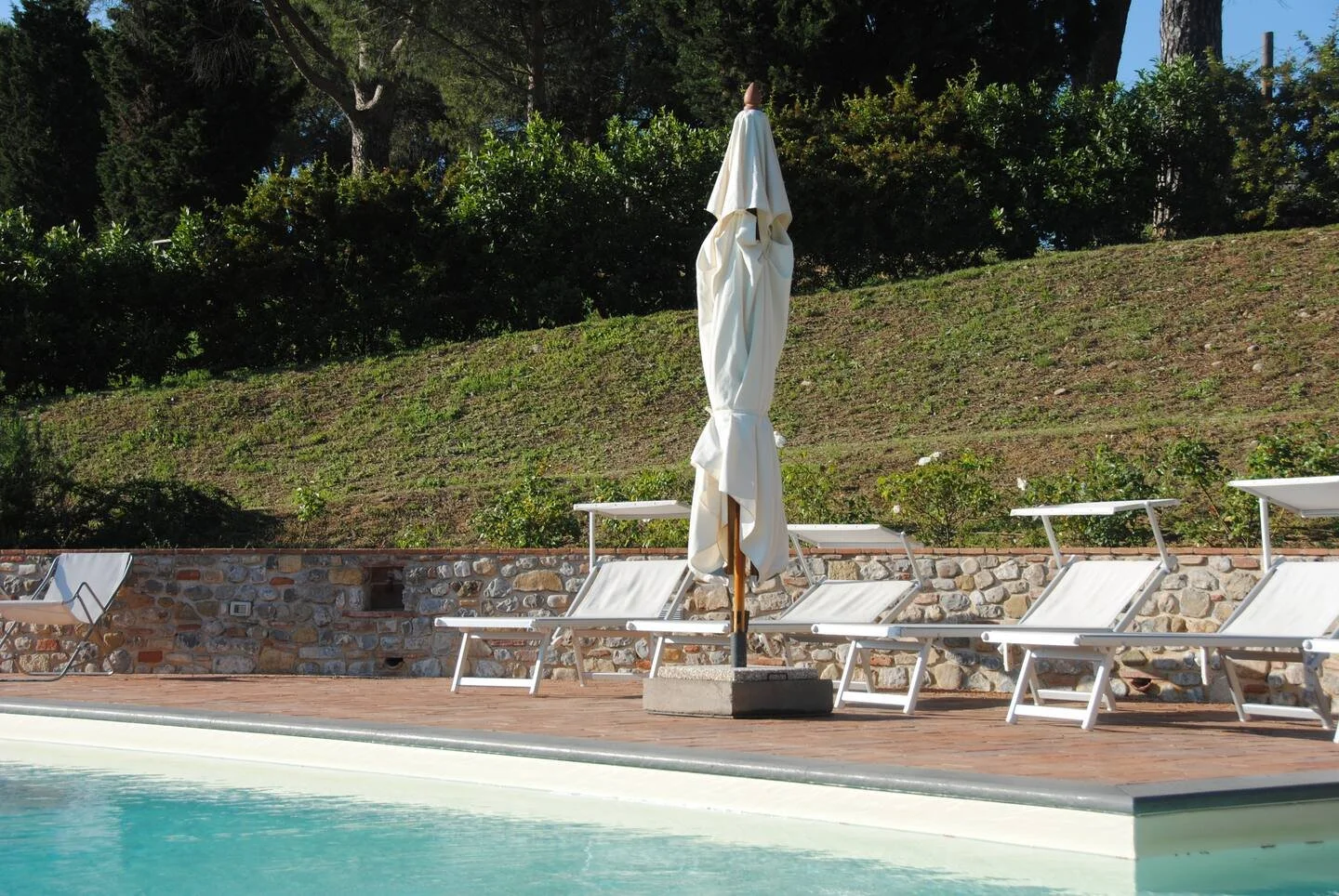 Empty poolside area with white lounge chairs and a closed umbrella, with a stone wall and green hillside in the background.