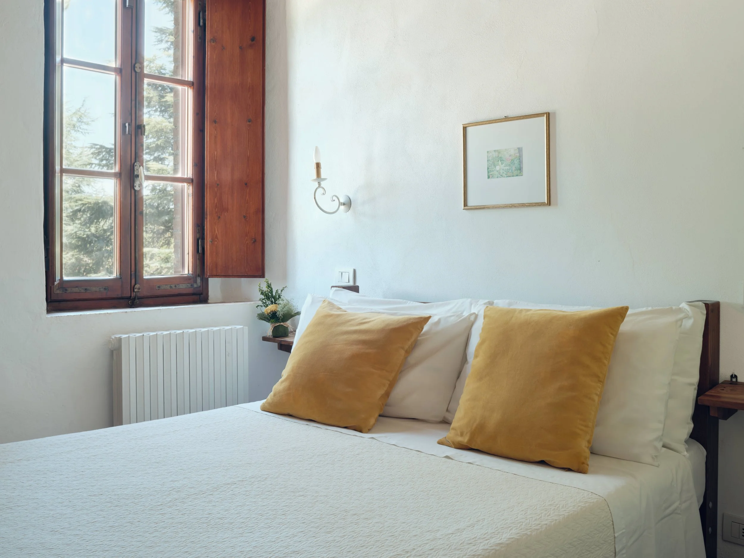 A bedroom corner with a large window, a bed with white bedding and two mustard-colored pillows, a small wooden bedside table with a vase of flowers, a white wall with a picture frame, a wall-mounted lamp, and a radiator below the window.