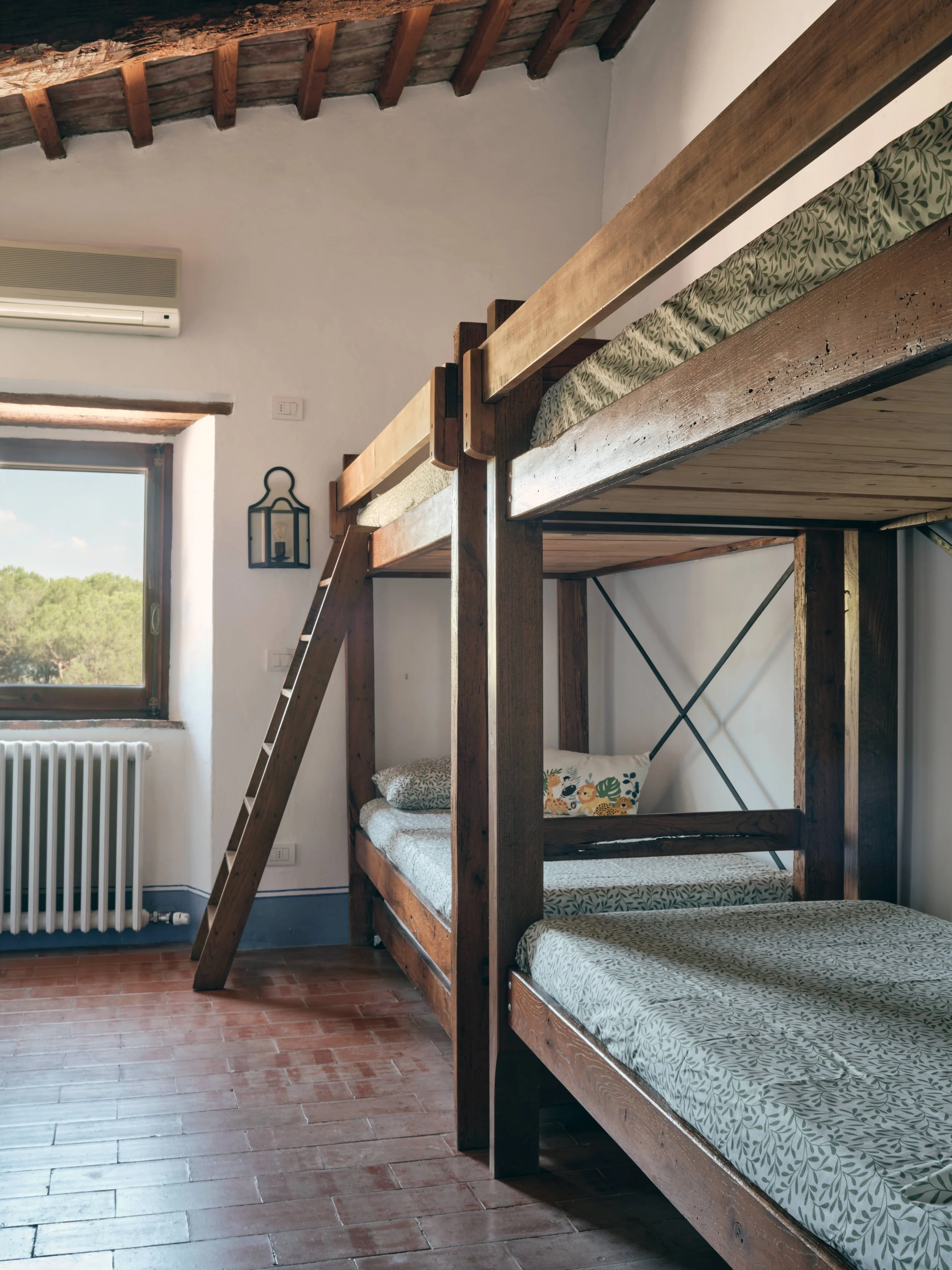 A wooden bunk bed with a ladder in a room with wooden floor and white walls. There is a window with a view of trees outside, an air conditioning unit on the wall, and a wall-mounted lantern-style light fixture. The mattresses have leafy-patterned bed