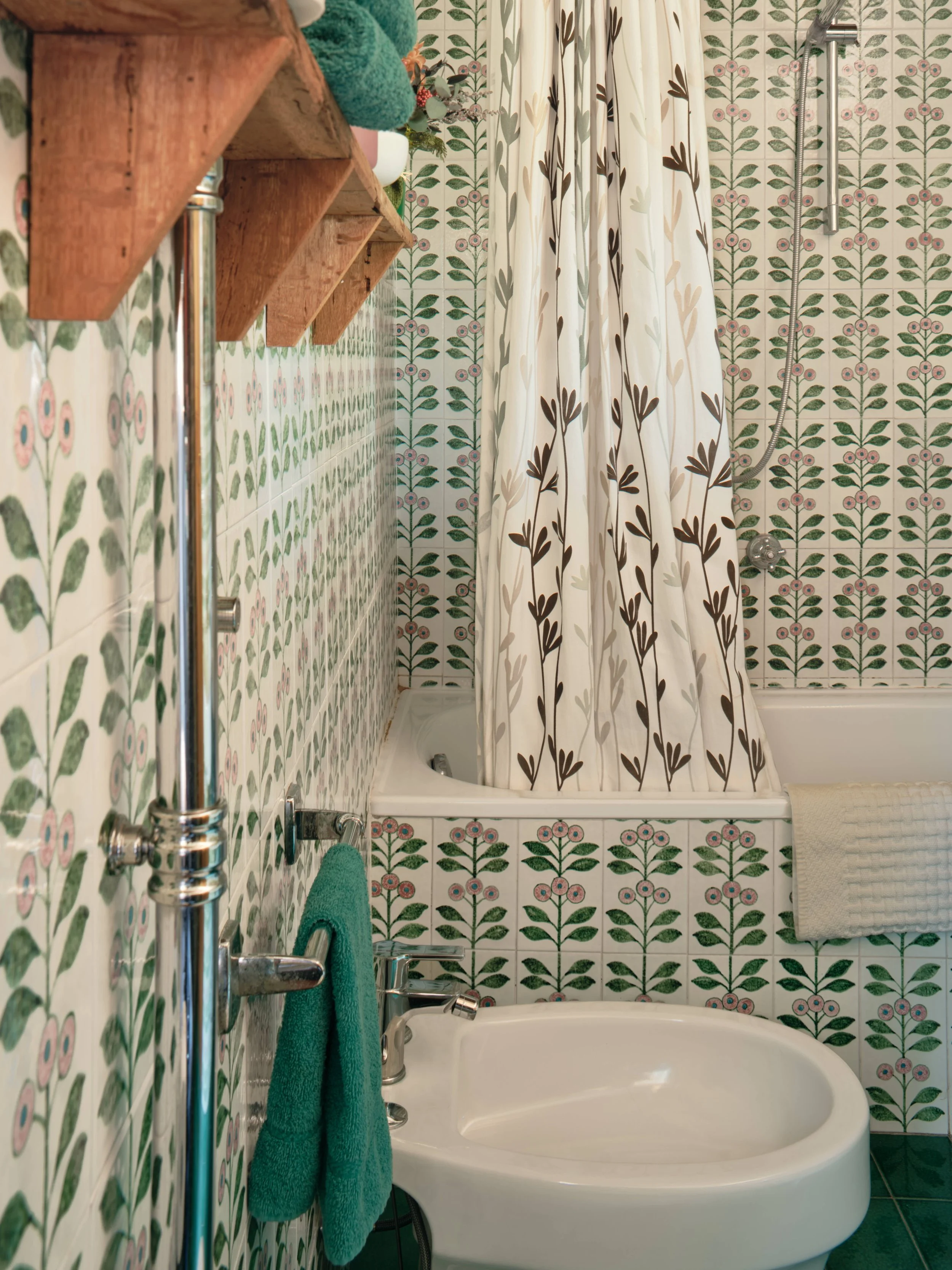 A bathroom with floral patterned tiles on the wall, featuring a bathtub with a showerhead and a curtain. A towel is hanging on a chrome towel bar, and a wooden shelf holds rolled towels and decorative items.