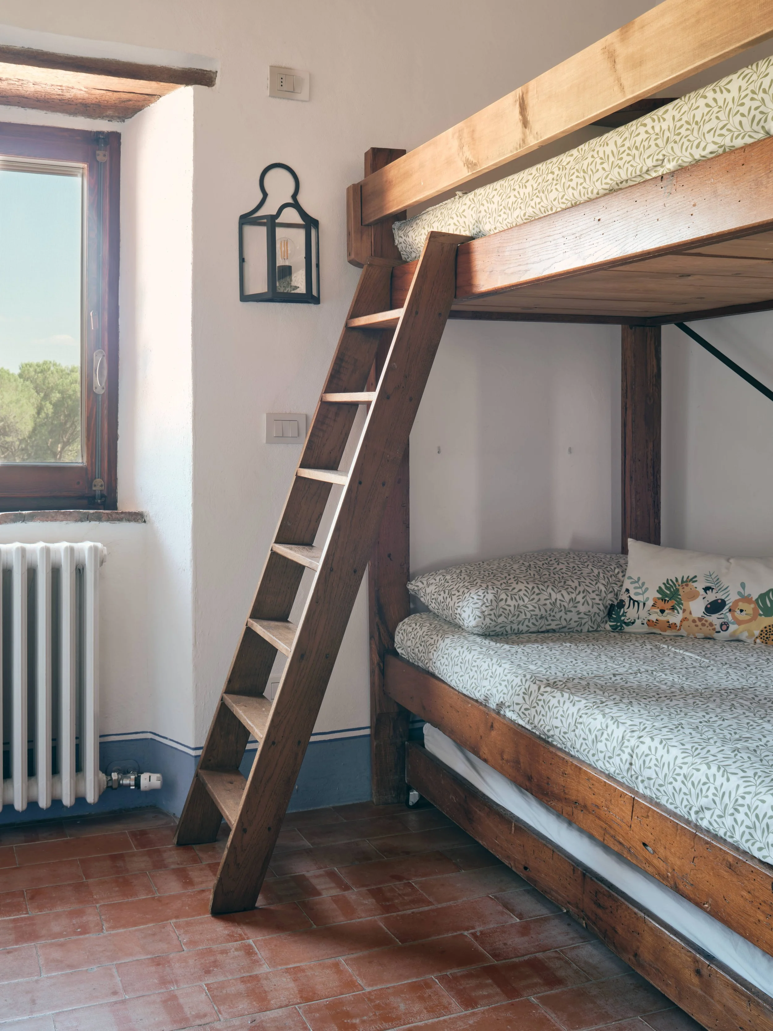 A bedroom with a wooden bunk bed, a ladder, a window, and a radiator. The bunk bed has a patterned bedsheet and decorative pillows with animal illustrations.