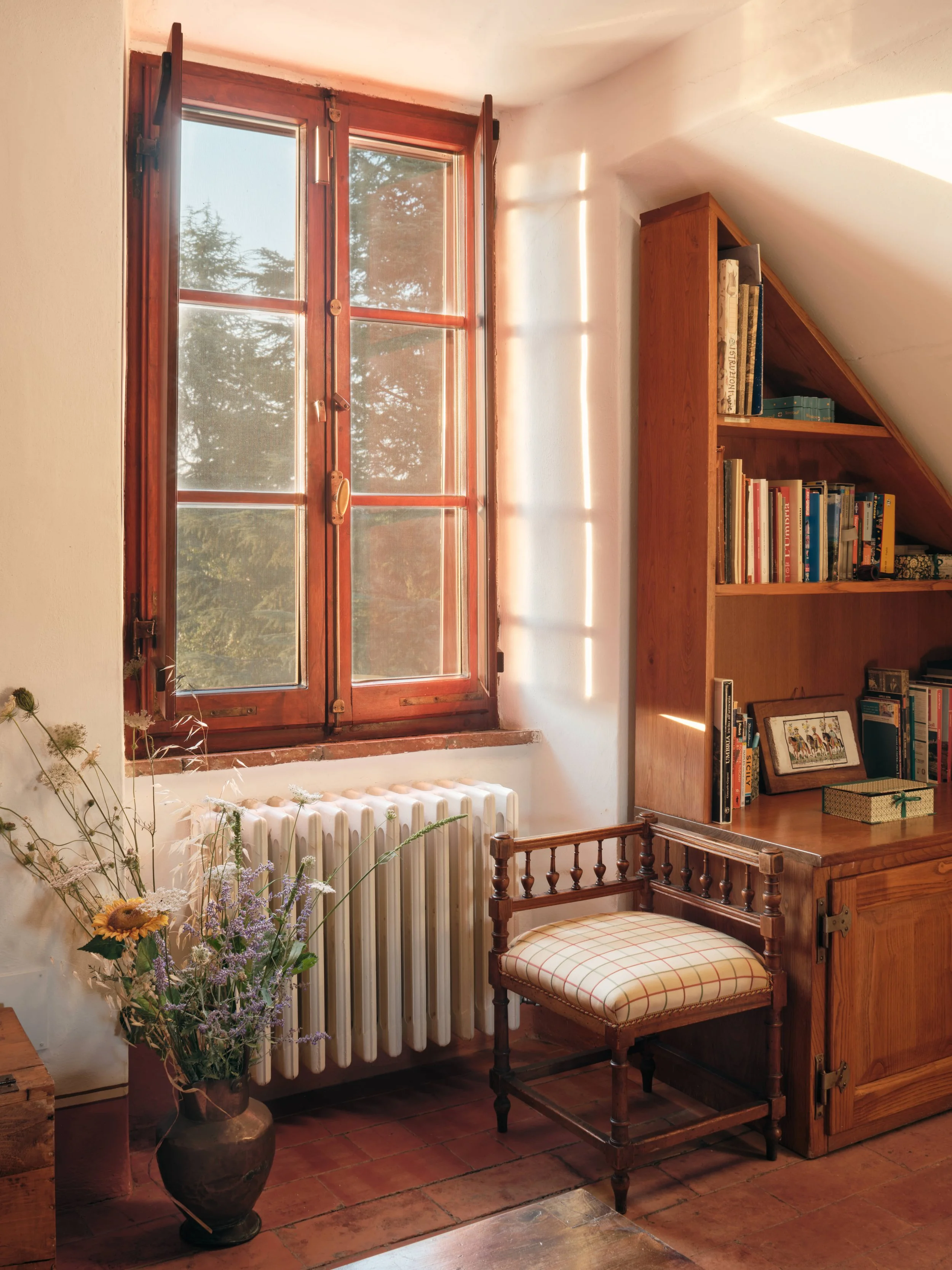 A cozy room with a wooden window letting in sunlight. A tall vase with dried and fresh flowers sits next to a white radiator. There is a wooden bookshelf filled with books and a framed picture. An upholstered chair with a checked cushion is in front 