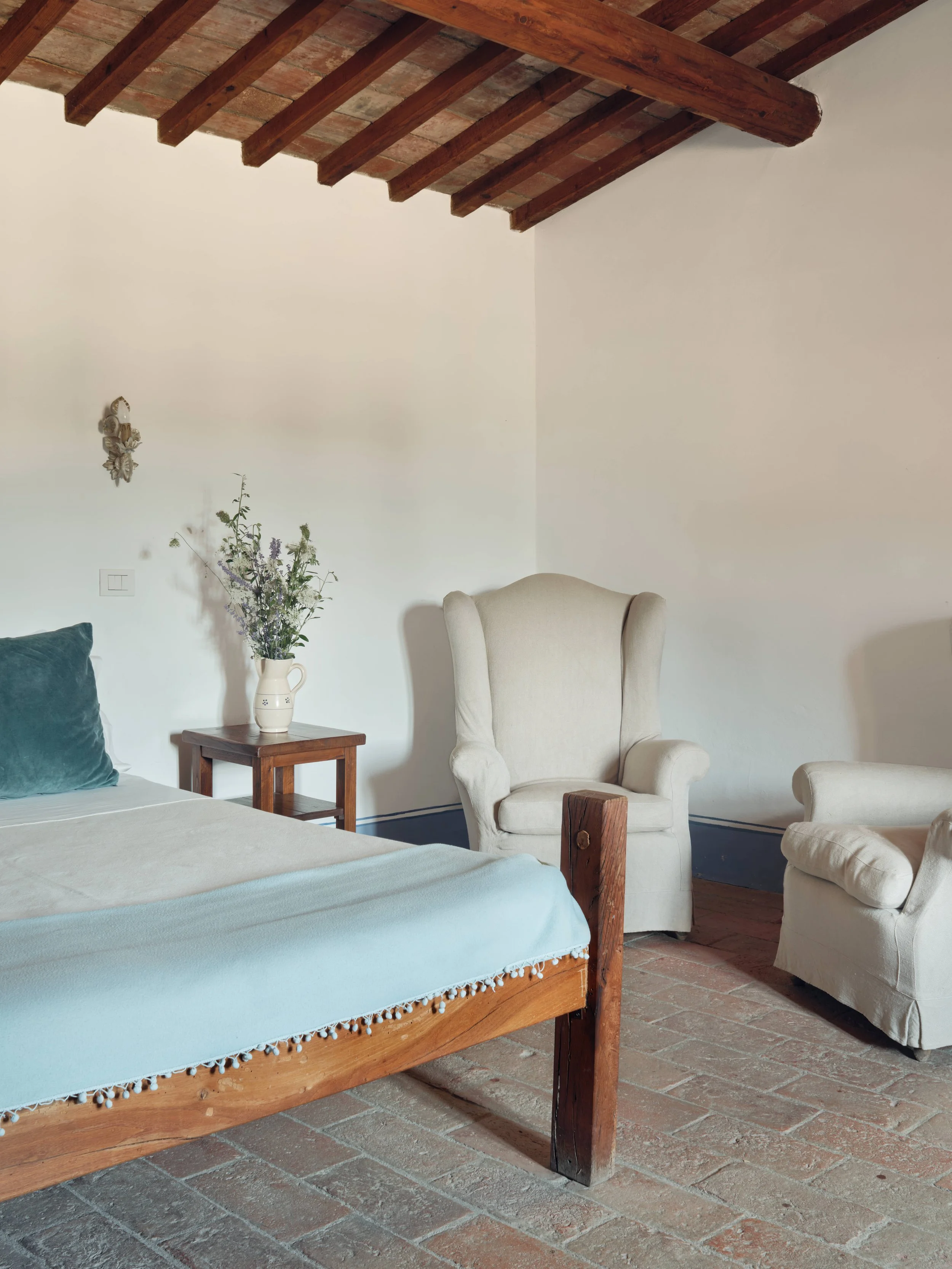 A cozy bedroom with a wooden bed frame, a white bedspread, a green pillow, a small wooden nightstand with a white pitcher flower vase, a beige armchair, and a white armchair, all set against a white wall with a decorative wall hanging.