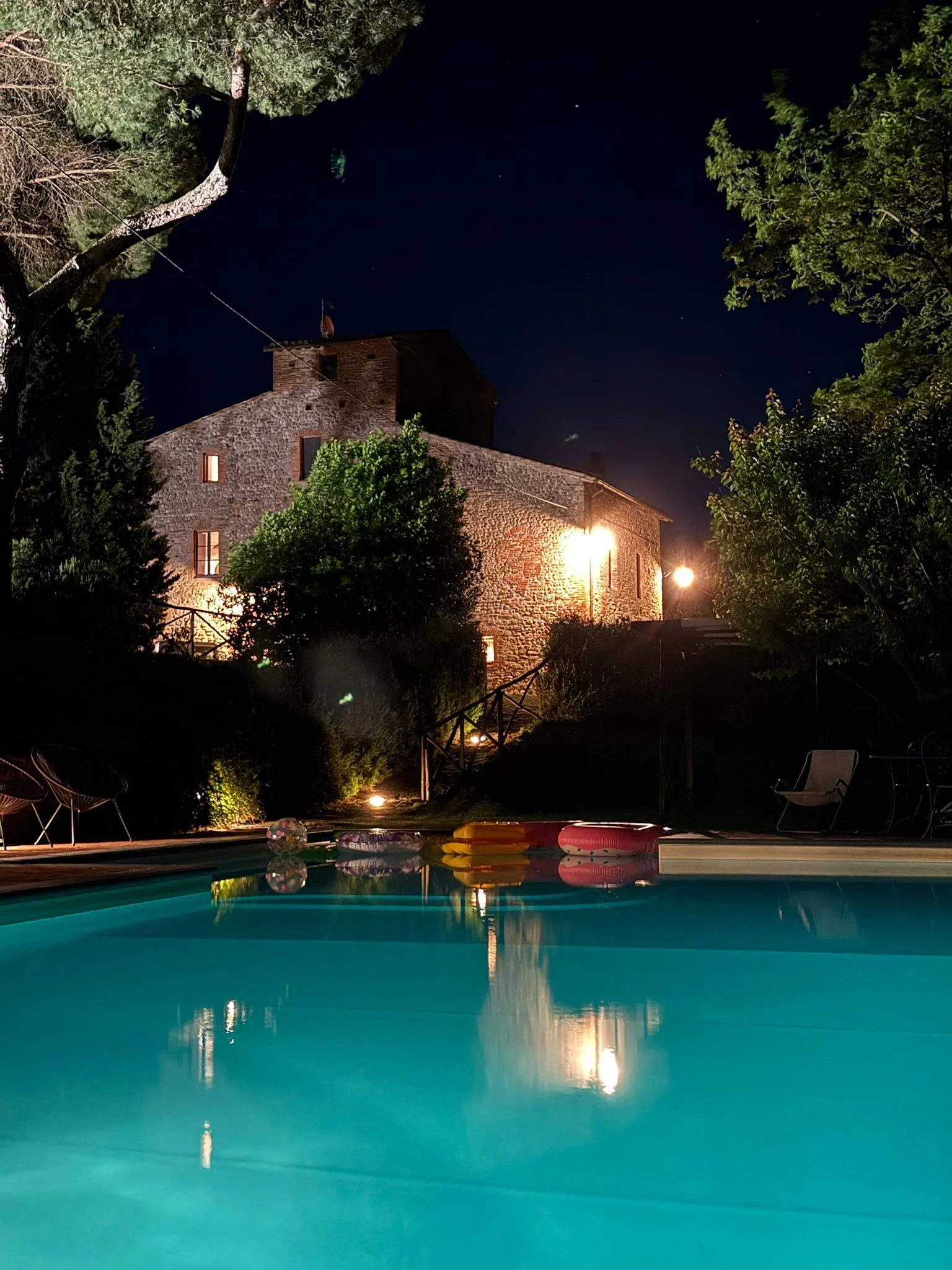 Nighttime view of a house with stone walls, illuminated by exterior lights, surrounded by trees, with stars visible in the dark sky; in the foreground, a swimming pool with a few pool floats reflected on the water's surface.