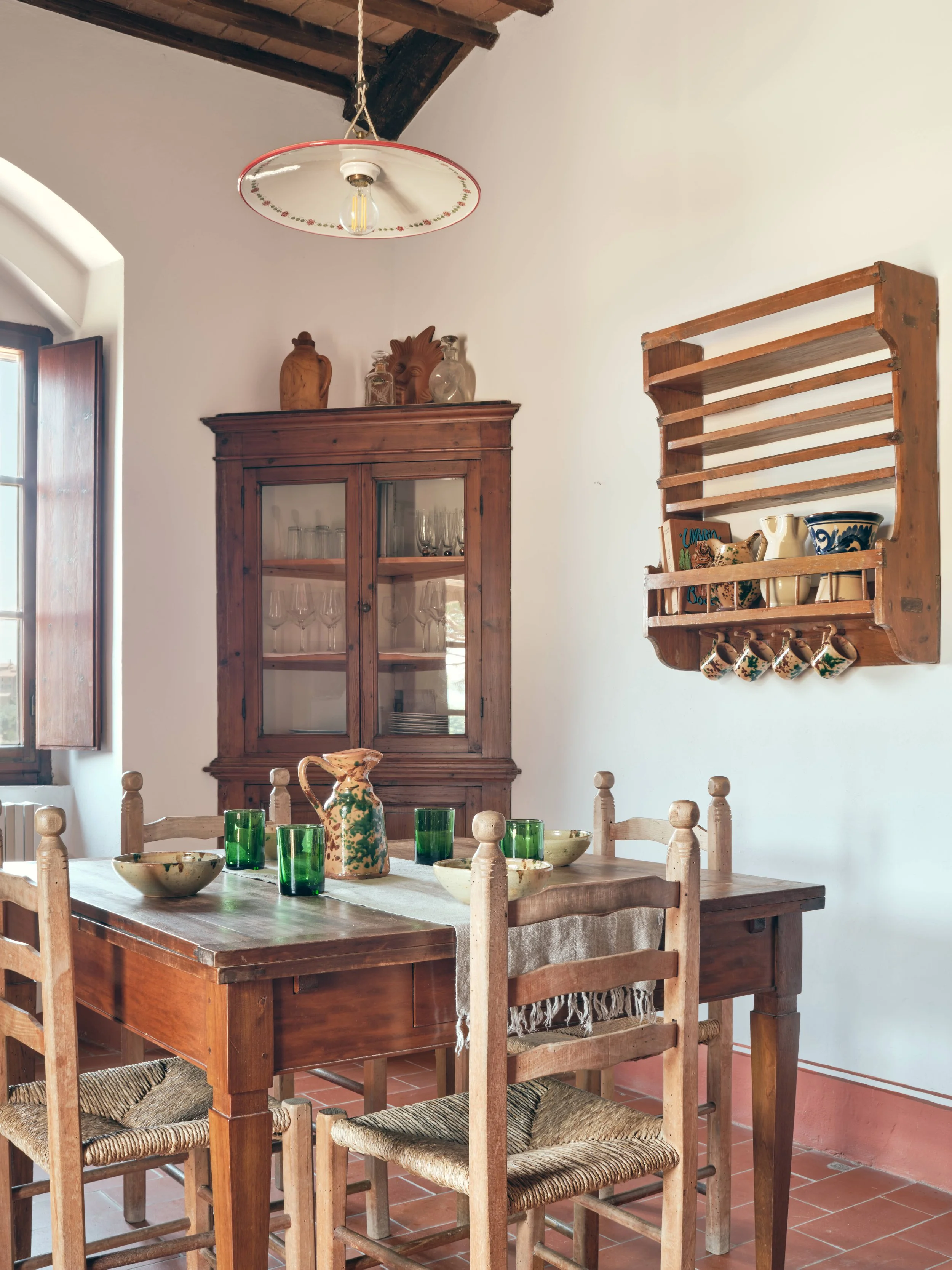 A rustic dining room with a wooden table set with bowls and green glasses, surrounded by wooden chairs with woven seats, a wooden cabinet with glass doors displaying glassware, and a wall-mounted wooden shelf holding ceramics and mugs. A window with 