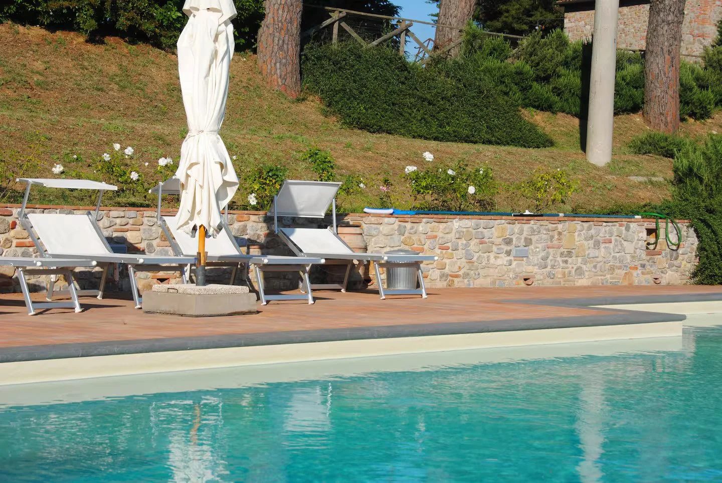 Empty poolside area with white lounge chairs, a closed white umbrella, and a stone wall background surrounded by greenery and trees.