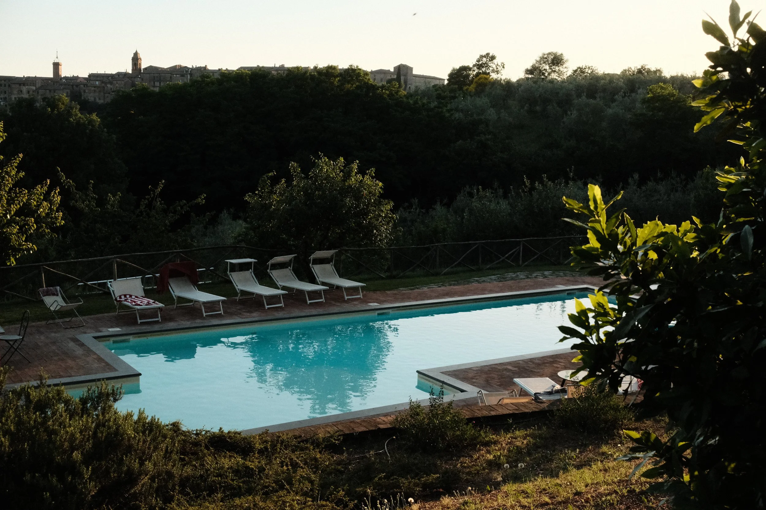 A swimming pool surrounded by lounge chairs, with a hillside town and green trees in the background, during sunset.