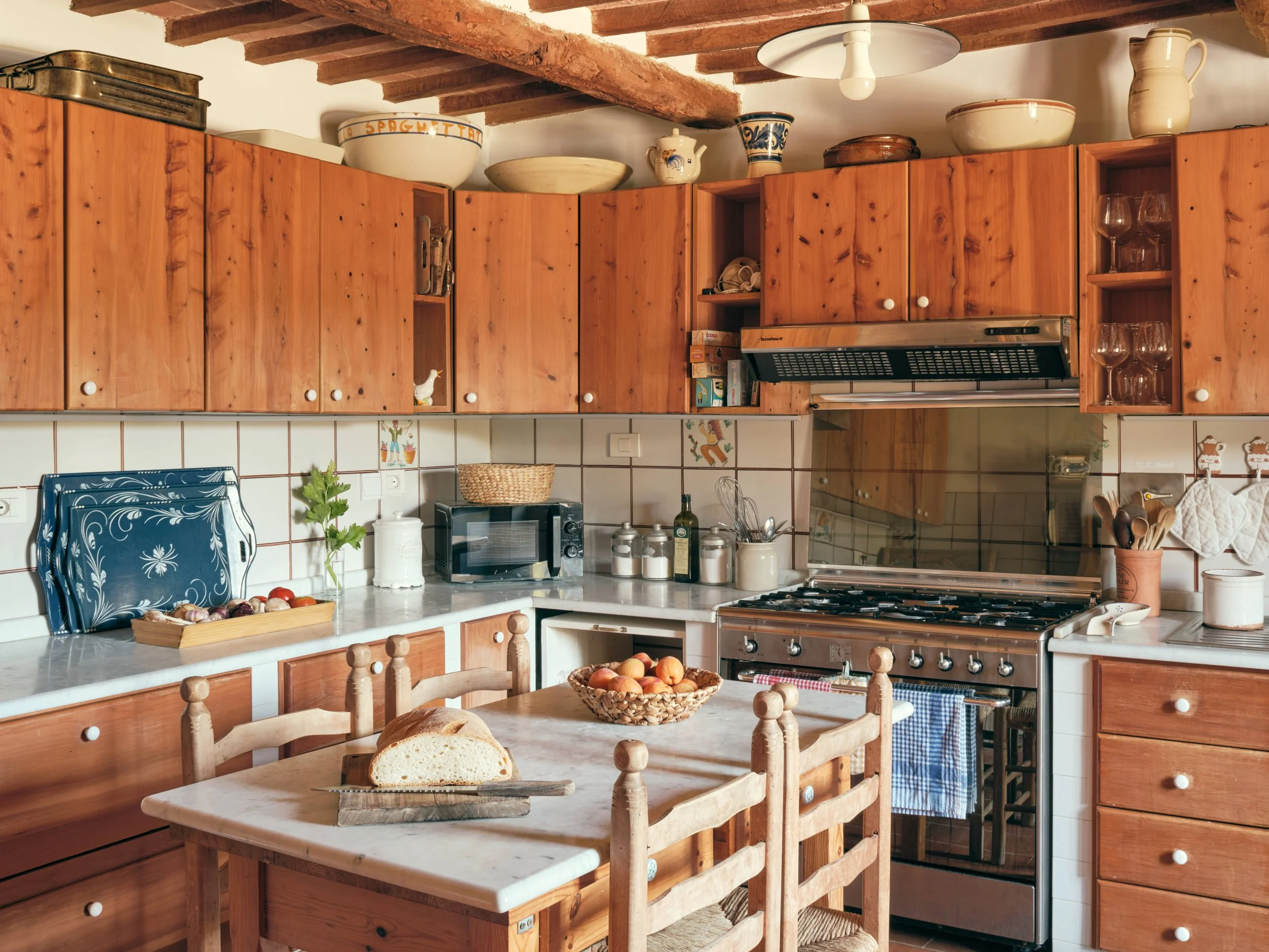 A cozy rustic kitchen with wooden cabinets and a marble-topped island. The shelves hold dishes, glassware, and kitchenware. There are fresh apples in a basket on the counter, and bread on the island. The ceiling features exposed wooden beams.