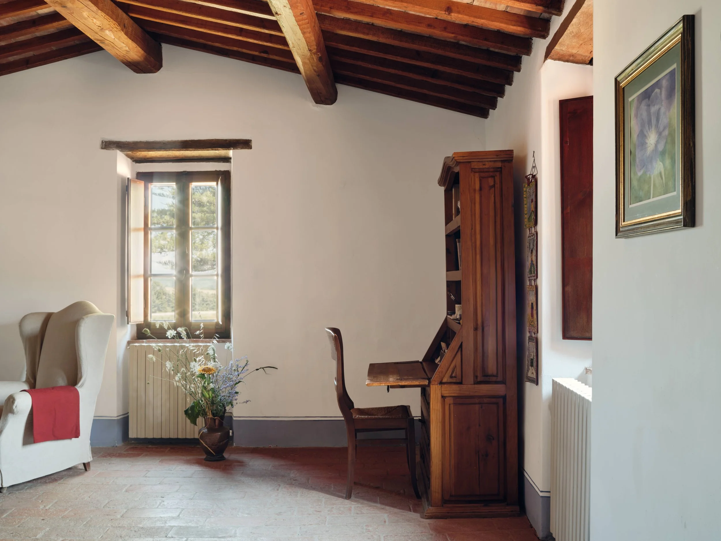 Interior of a cozy room with white walls and a wooden ceiling with exposed beams. A window lets in natural light, revealing trees outside. There's a white armchair with a red throw blanket, a floral arrangement, a wooden cabinet, and framed artwork o