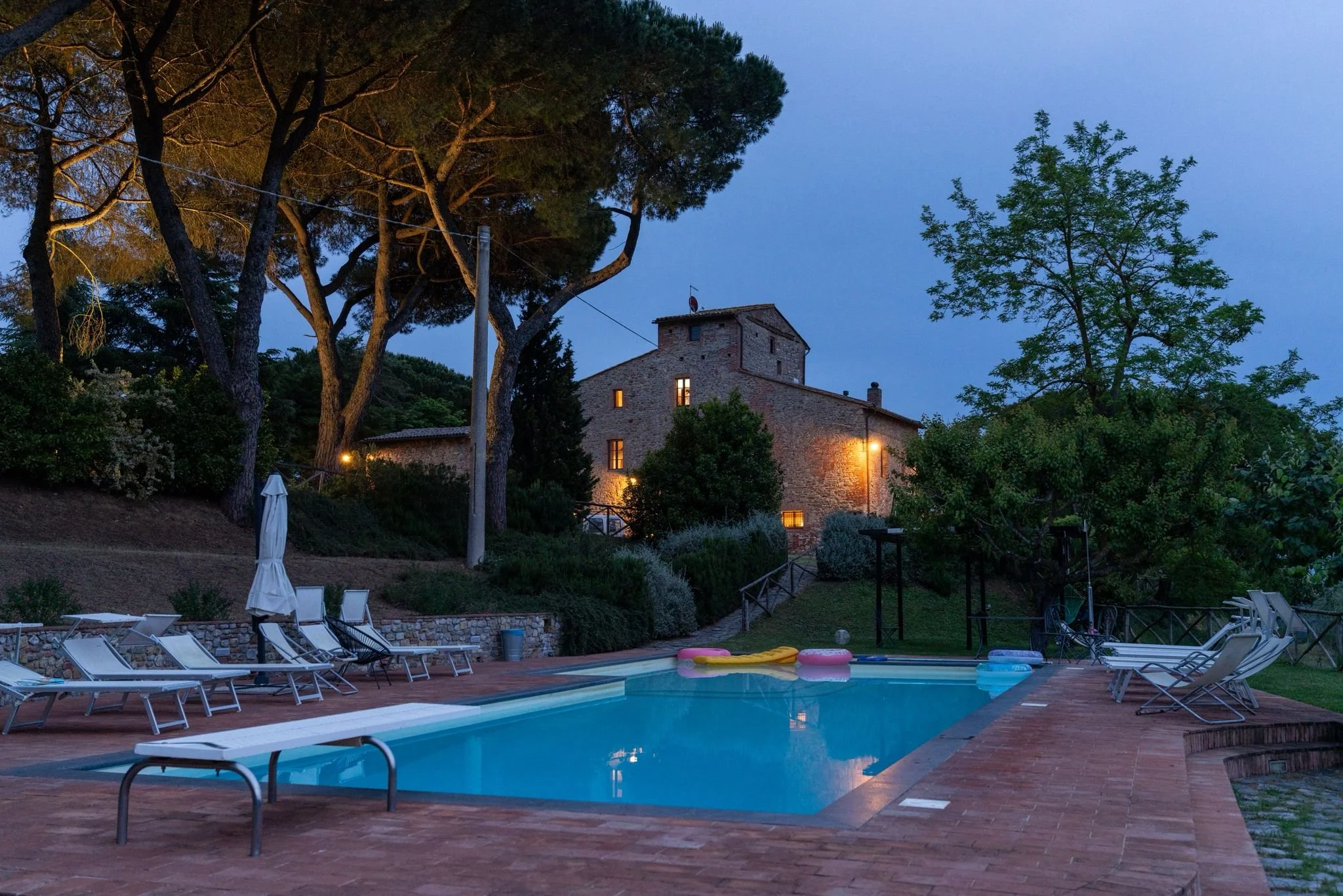 A backyard pool area in the evening with pool chairs, water toys, and an umbrella. In the background, there is a stone house surrounded by tall trees, with outdoor lights illuminating the scene.