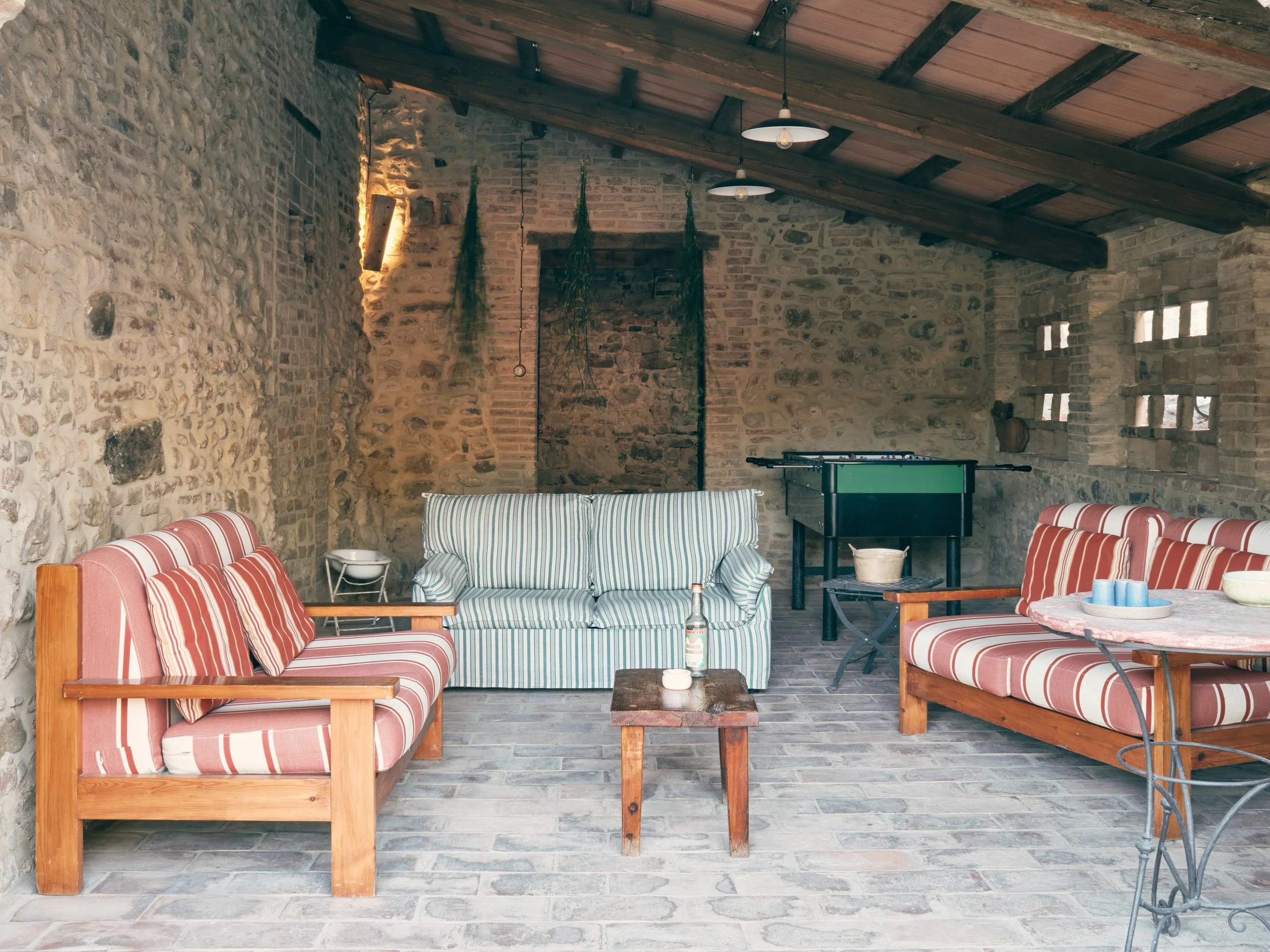 A cozy indoor space with exposed brick and stone walls and a wooden ceiling, furnished with striped and solid-colored sofas, wooden tables, and a foosball table, decorated with hanging plants and warm lighting.