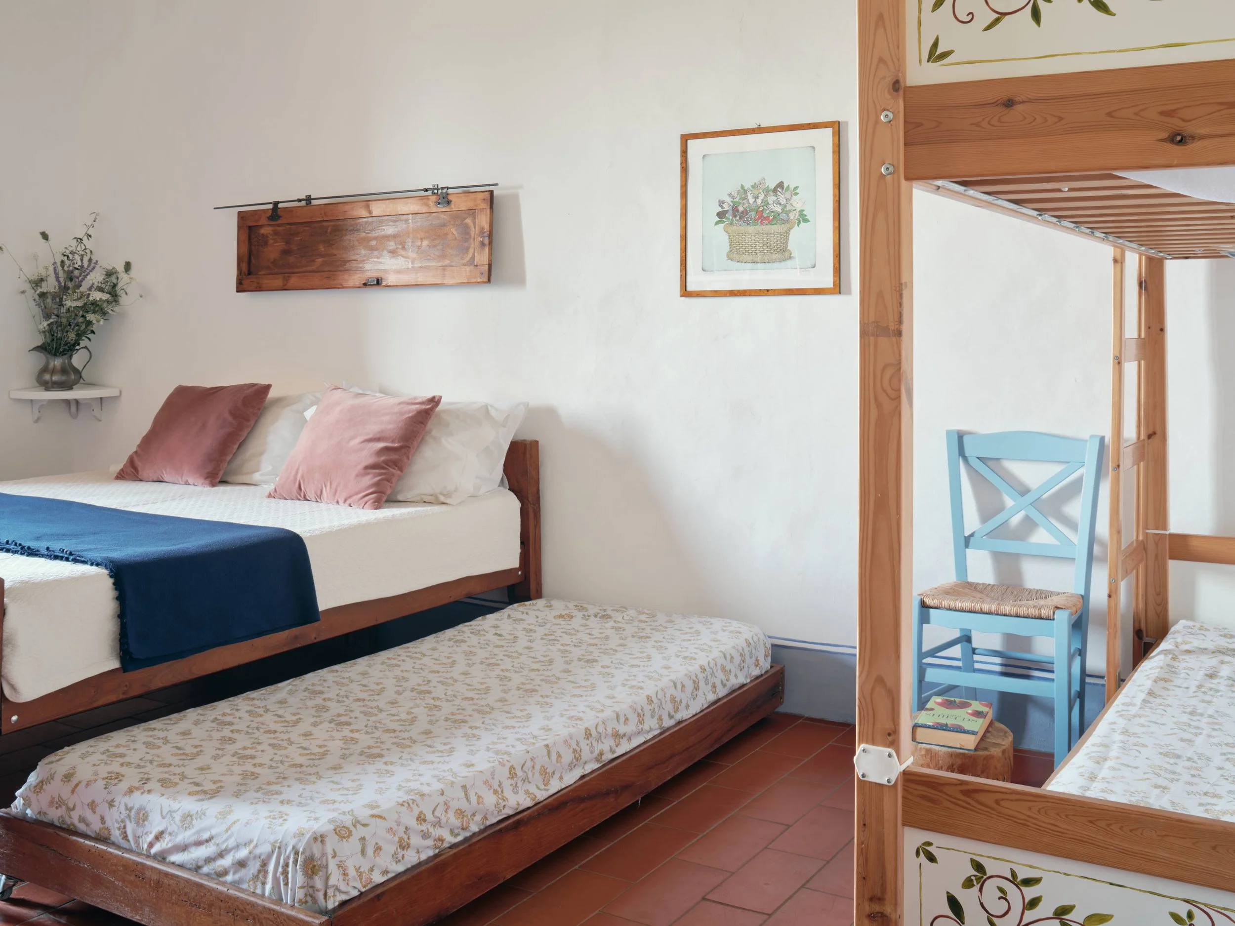 Room with a bed that has pink pillows, a framed flower basket painting on the wall, a wooden shelf with a vase and flowers, and a blue chair beside a wooden structure.