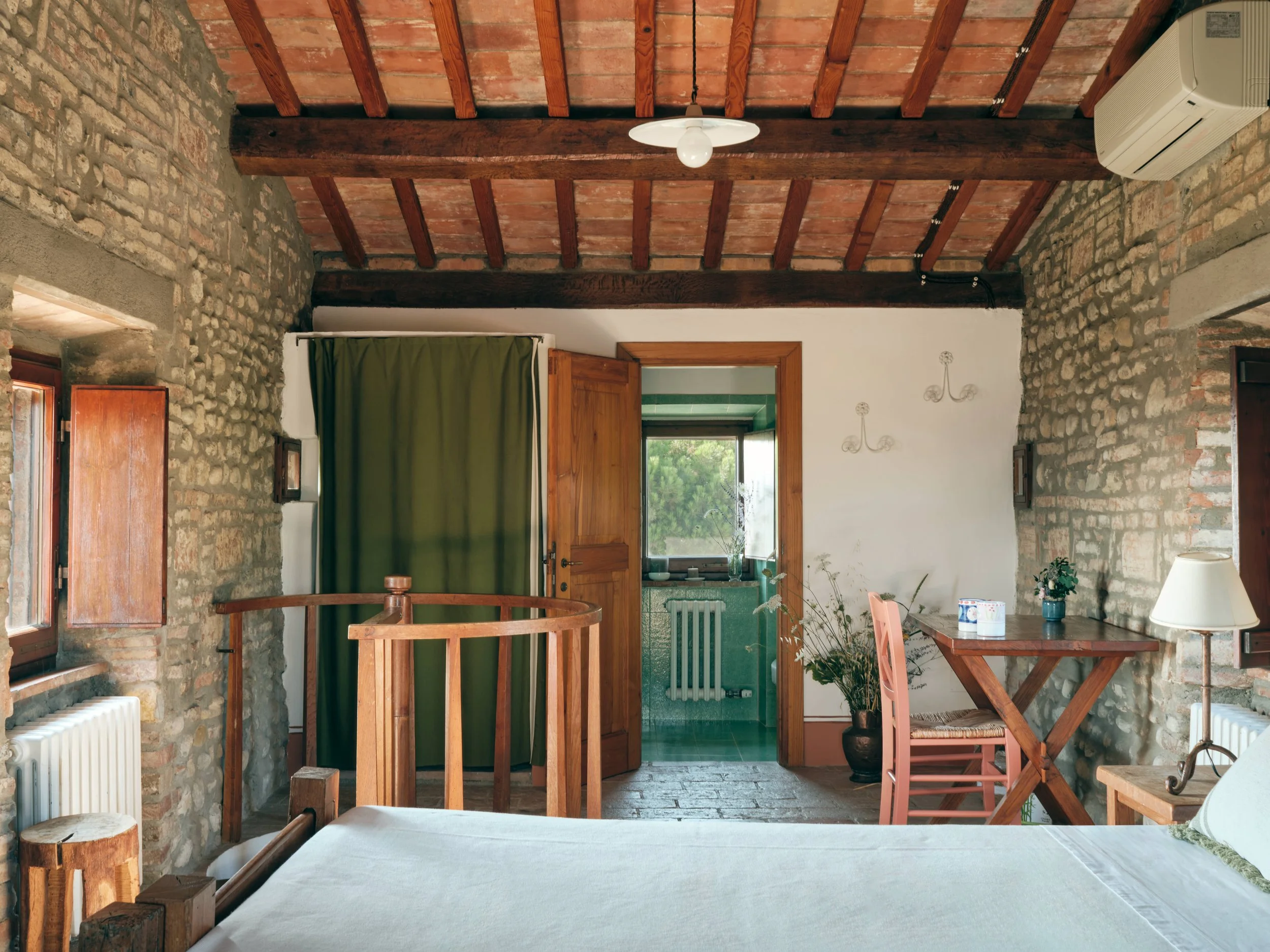 Rustic dining room with brick and stone walls, wooden ceiling beams, green curtain, wooden window and door, and a dining table with a white tablecloth. There is also a potted plant and a small table with a lamp.