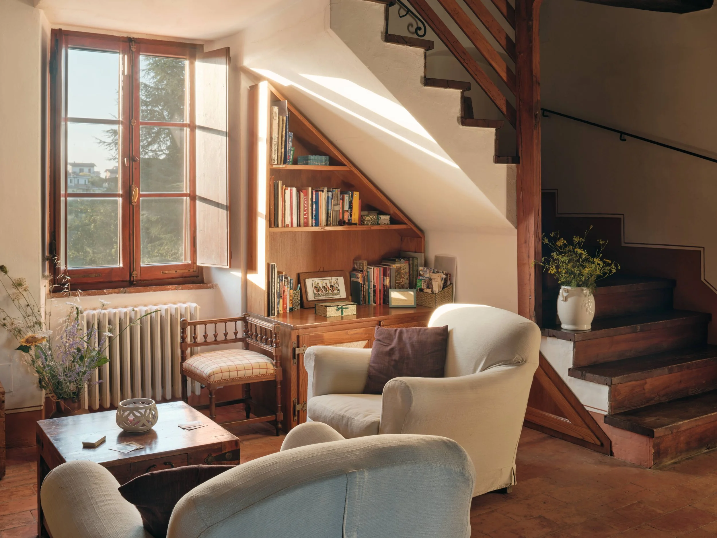Living room with a wooden staircase, a window letting in natural light, a bookshelf, a beige armchair, a wooden coffee table, and a vase with flowers.