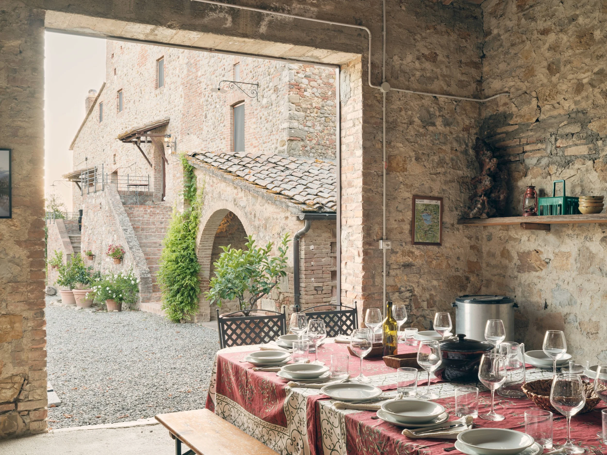 Indoor dining area with a rustic stone wall, set for a meal with glasses, plates, and a bottle of olive oil, overlooking an outdoor courtyard with potted plants and stone buildings.