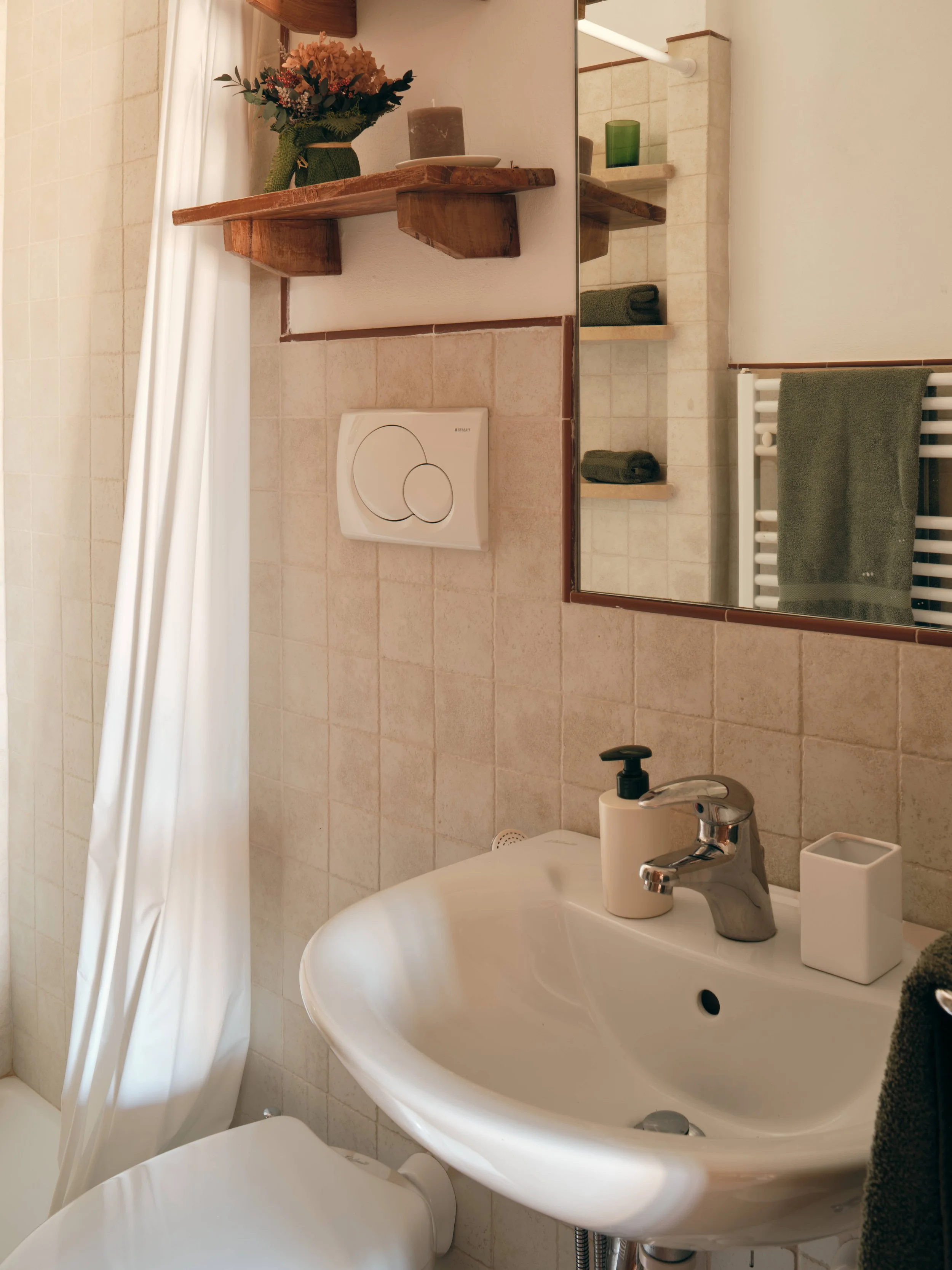 A bathroom with a sink, mirror, and toilet, featuring a beige tiled wall, a green towel on a white radiator, a black bottle, a white container, and a partially visible shower curtain with a wooden shelf holding a vase with flowers and candles.