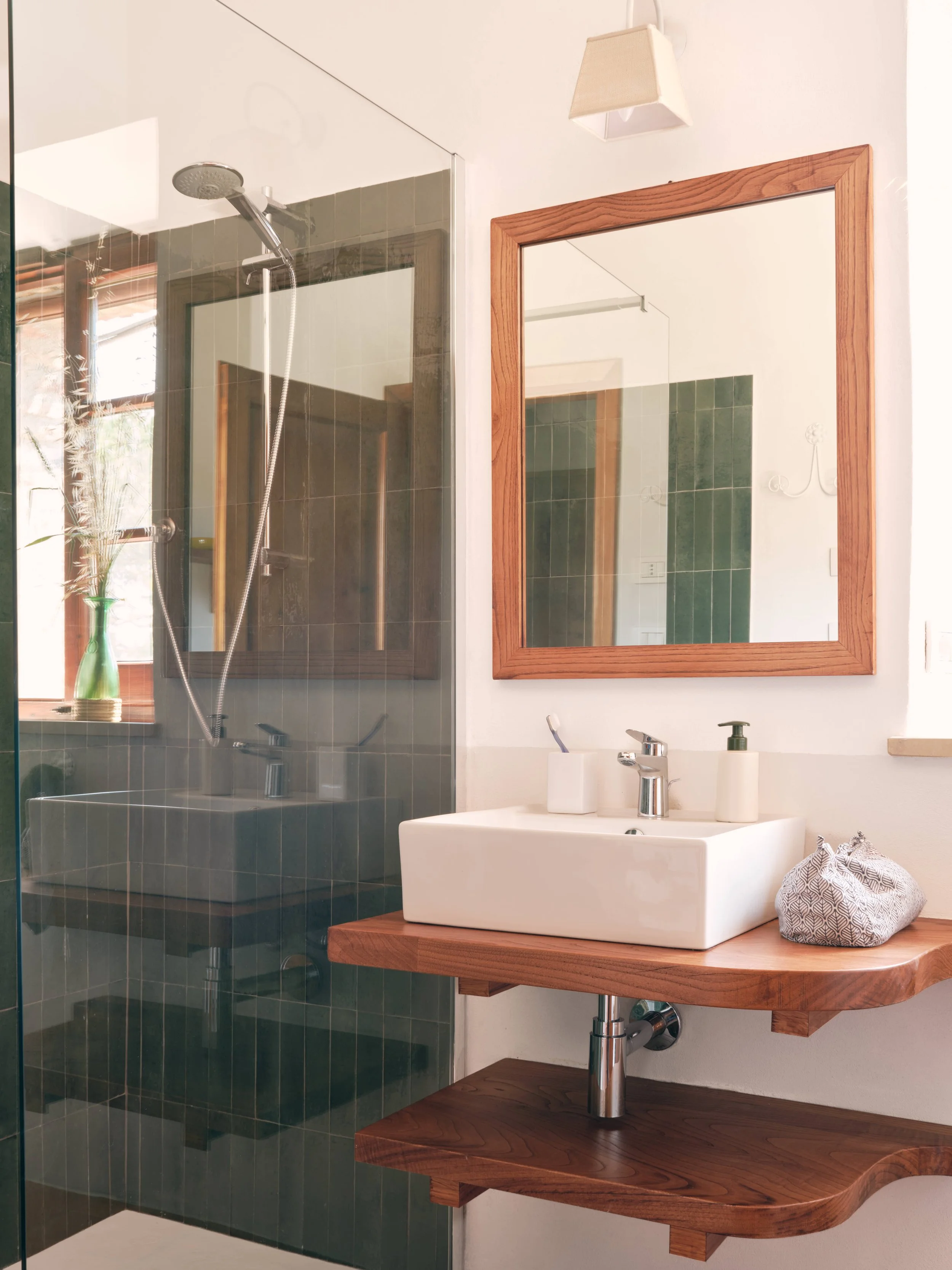 Bathroom with a white ceramic sink on a wooden countertop, a wooden framed mirror, a glass shower enclosure with a showerhead, a green vase with dried plants, and a wall-mounted light fixture.