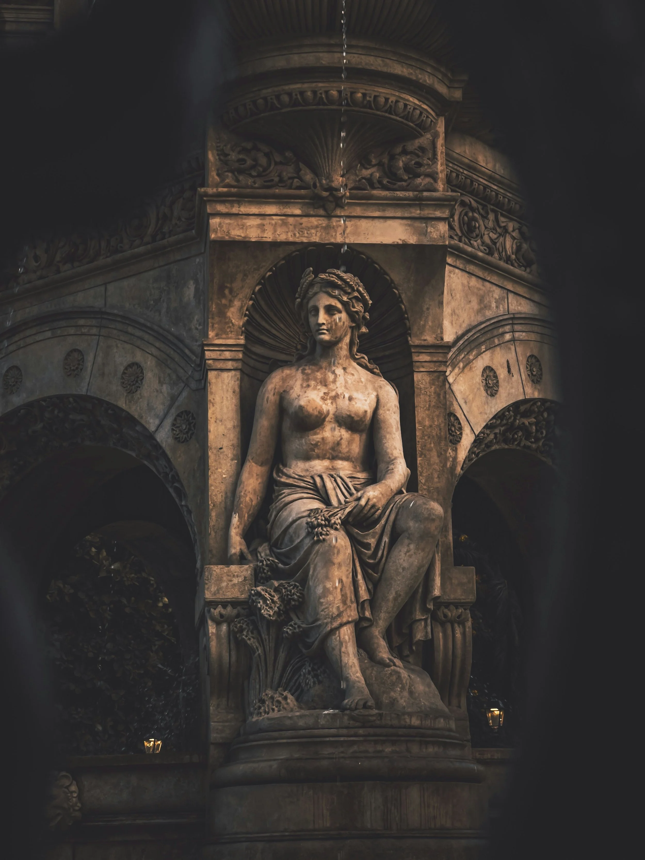 A classical marble statue of a seated woman with a crown of leaves, set in an ornate architectural niche with decorative carvings and a fountain with water flowing from the top.
