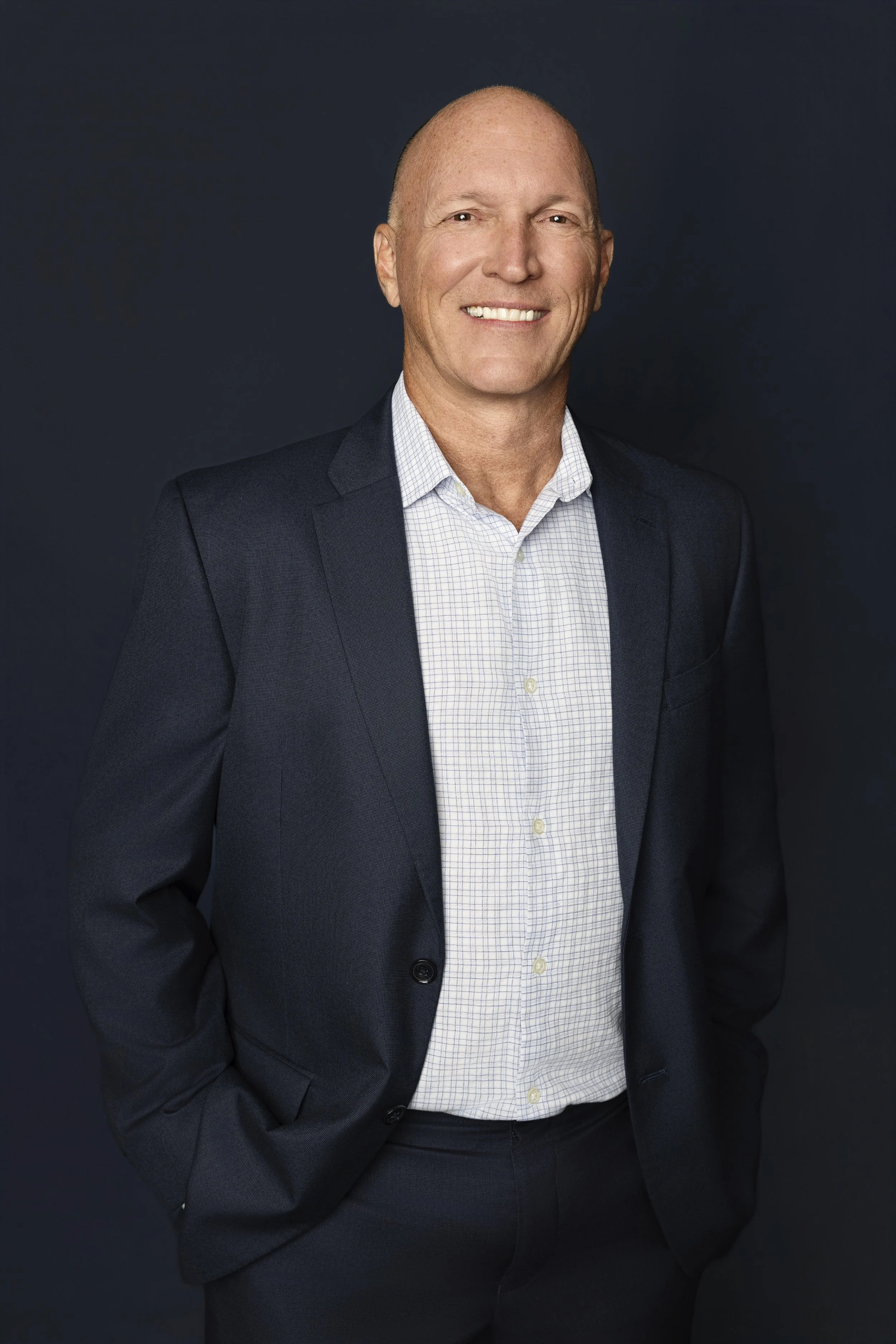 A professional headshot of a smiling bald man wearing a dark suit jacket and a white checkered shirt against a dark background.