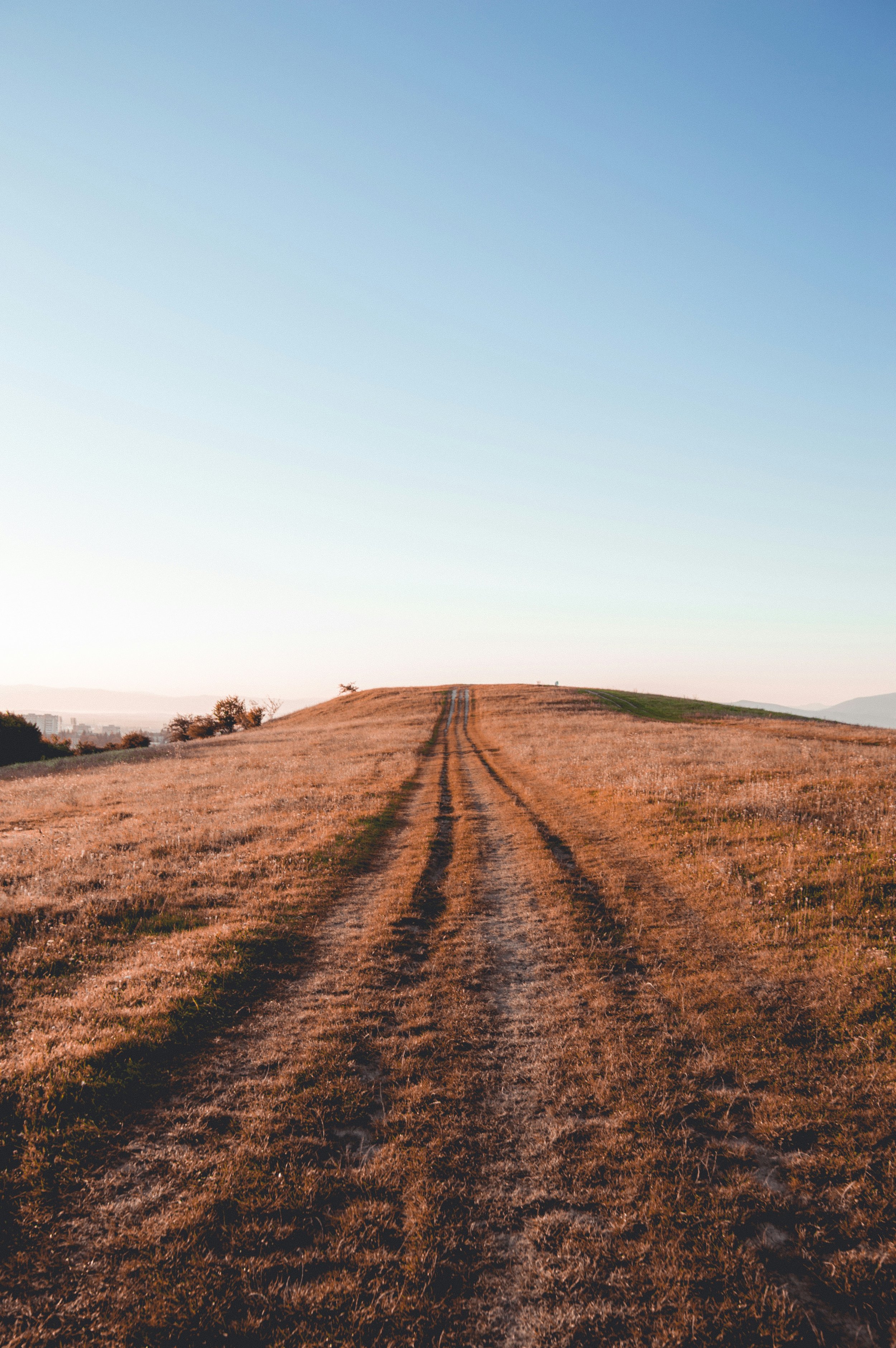 Dirt road running across grassy field, leading up a small hill with a clear sky in the background.