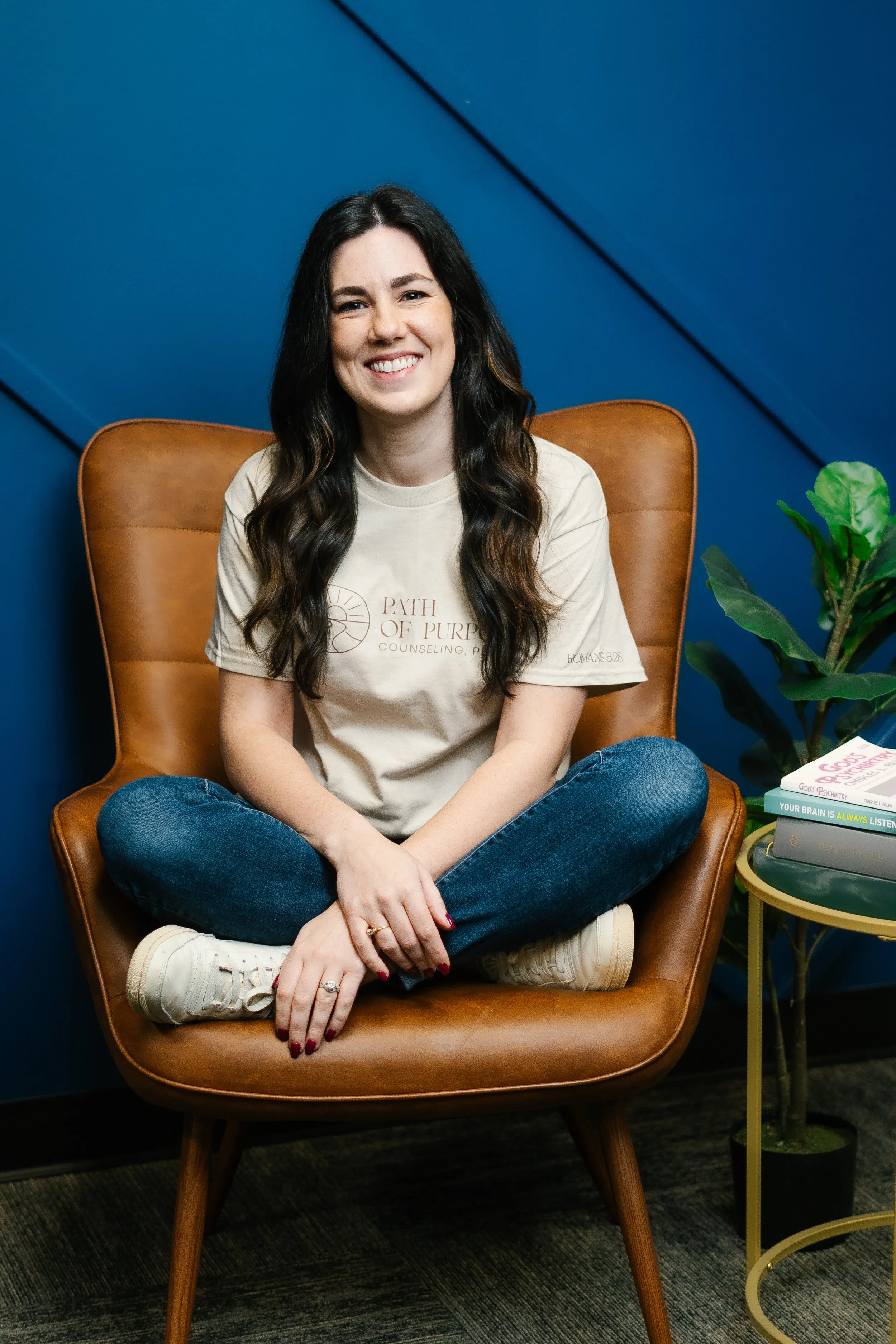 A young woman with long dark hair, sitting cross-legged in a brown armchair, smiling at the camera. She is wearing a beige t-shirt with text and jeans. Next to her is a small yellow side table with stacks of books and a green potted plant, against a blue wall.