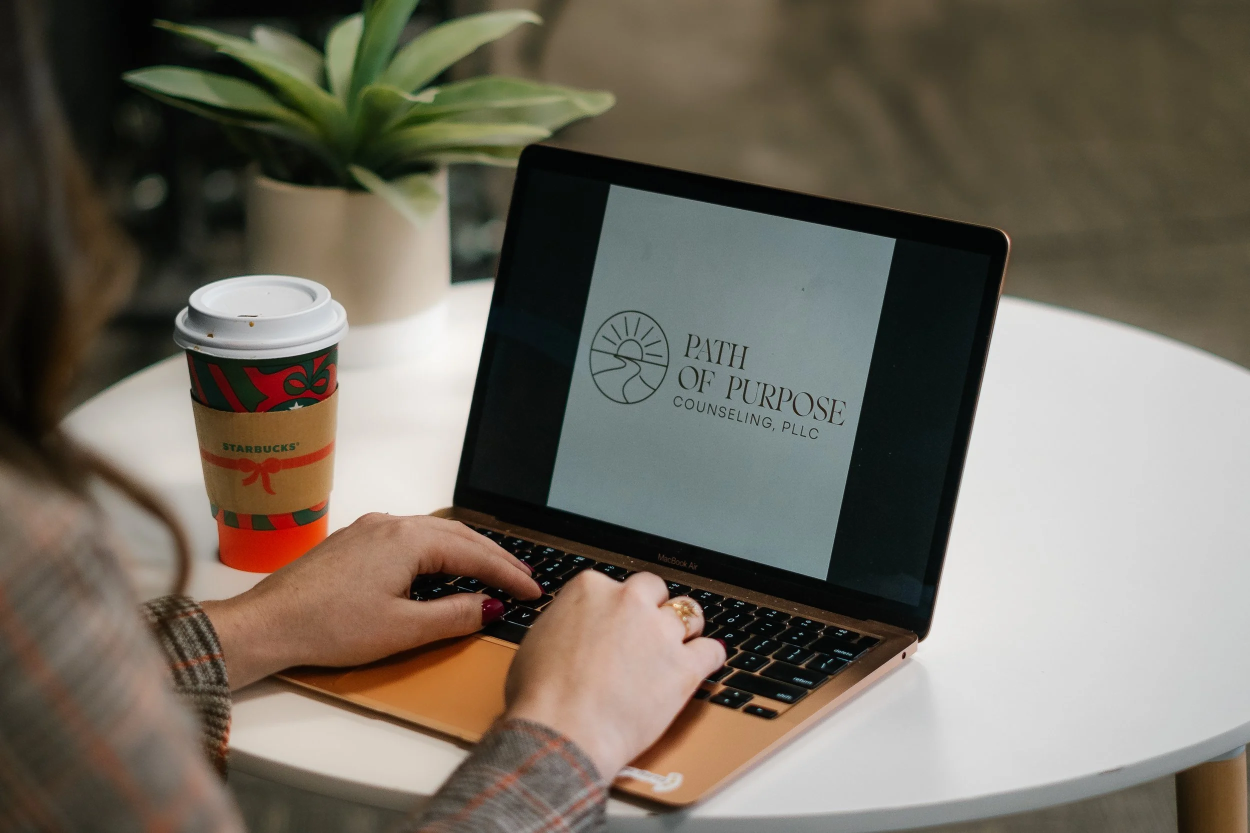 A person with painted nails using a MacBook Air laptop, with a Starbucks coffee cup and a potted plant in the background.