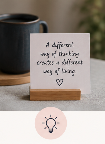 A white card with a motivational quote on it, placed in a wooden holder on a table next to a black coffee mug. The quote reads, "A different way of thinking creates a different way of living" with a small heart symbol underneath.