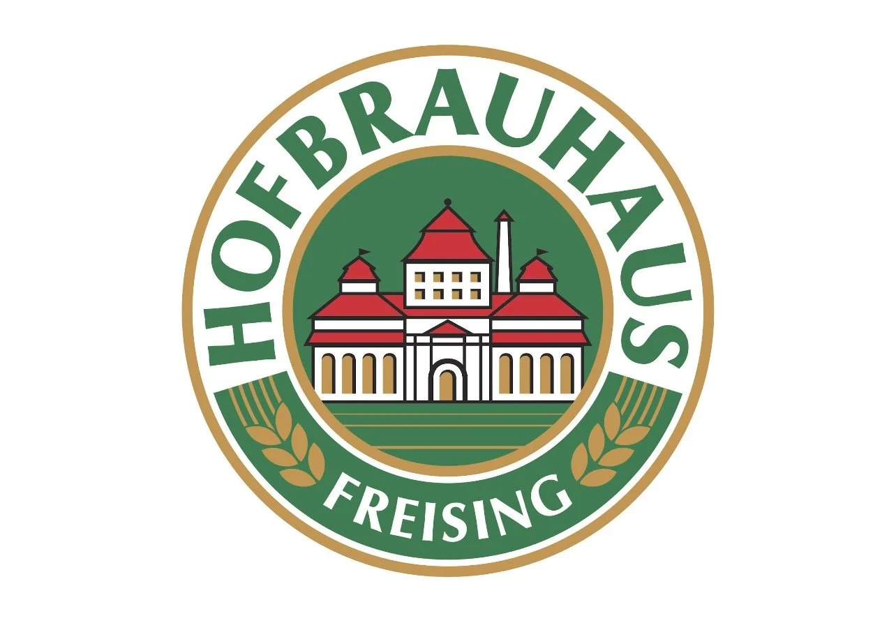 Logo of the House of Brauhaus Freising, featuring a historic building with red roofs and a chimney, surrounded by a green circle and gold laurel branches.