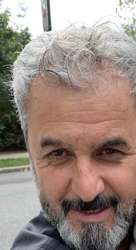 Close-up of a smiling man with gray hair, beard, and mustache, outdoor setting with trees and street in the background.