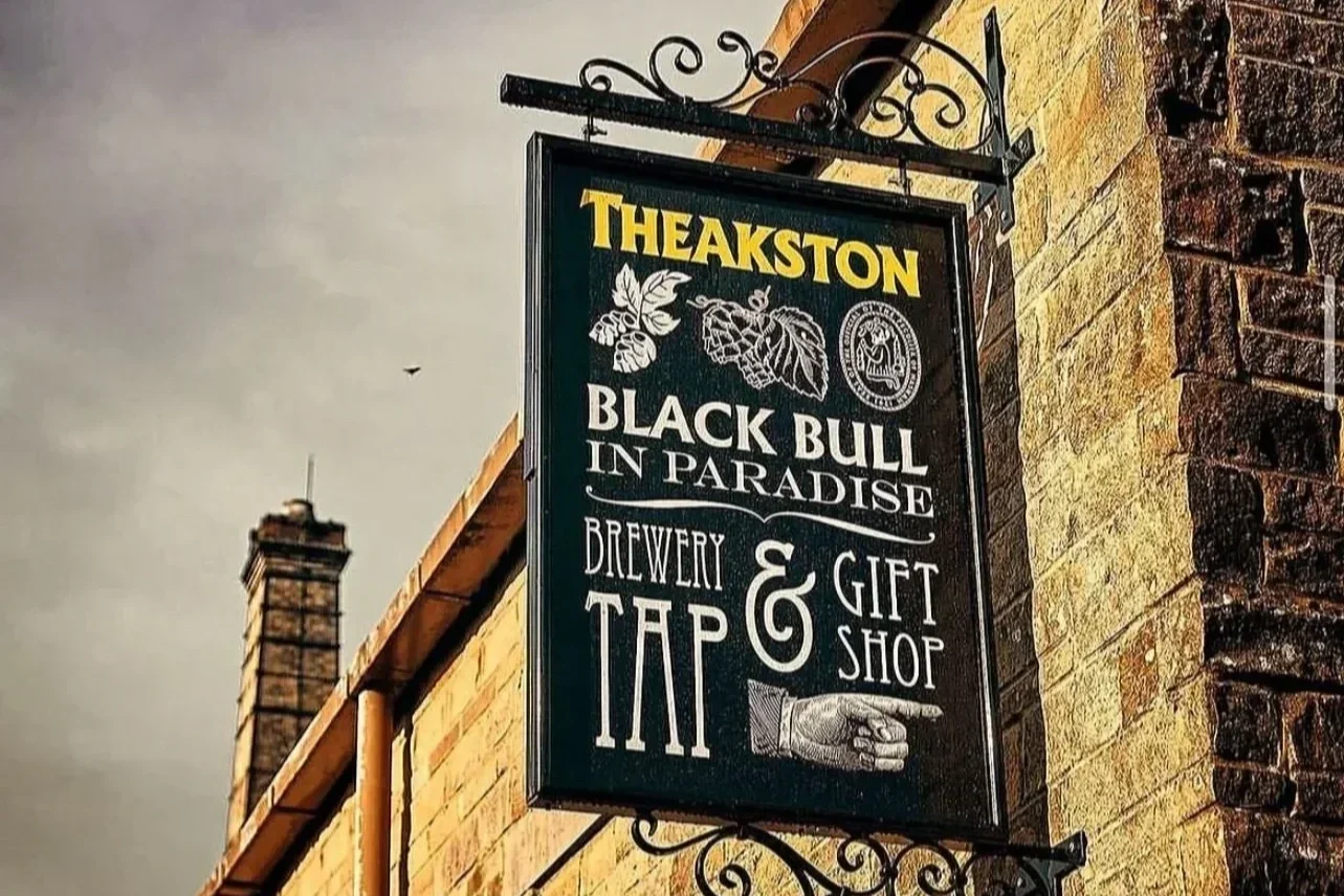 Black metal sign hanging on a brick building for Theakston brewery, advertising Black Bull in Paradise, a brew for tee, gift shop, with illustrations of hops, leaves, and a hand pointing right.