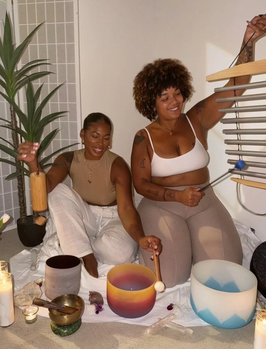 Two women practicing sound healing with crystal bowls, singing and smiling in a cozy indoor setting with candles, plants, and a white cloth on the floor.