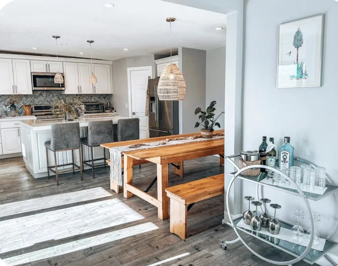Open-concept kitchen and dining area with white cabinets, stainless steel appliances, and a wooden dining table with benches and chairs. Decor includes potted plants, framed art, and a bar cart with bottles and glassware. Natural light casts shadows on the wooden floor.