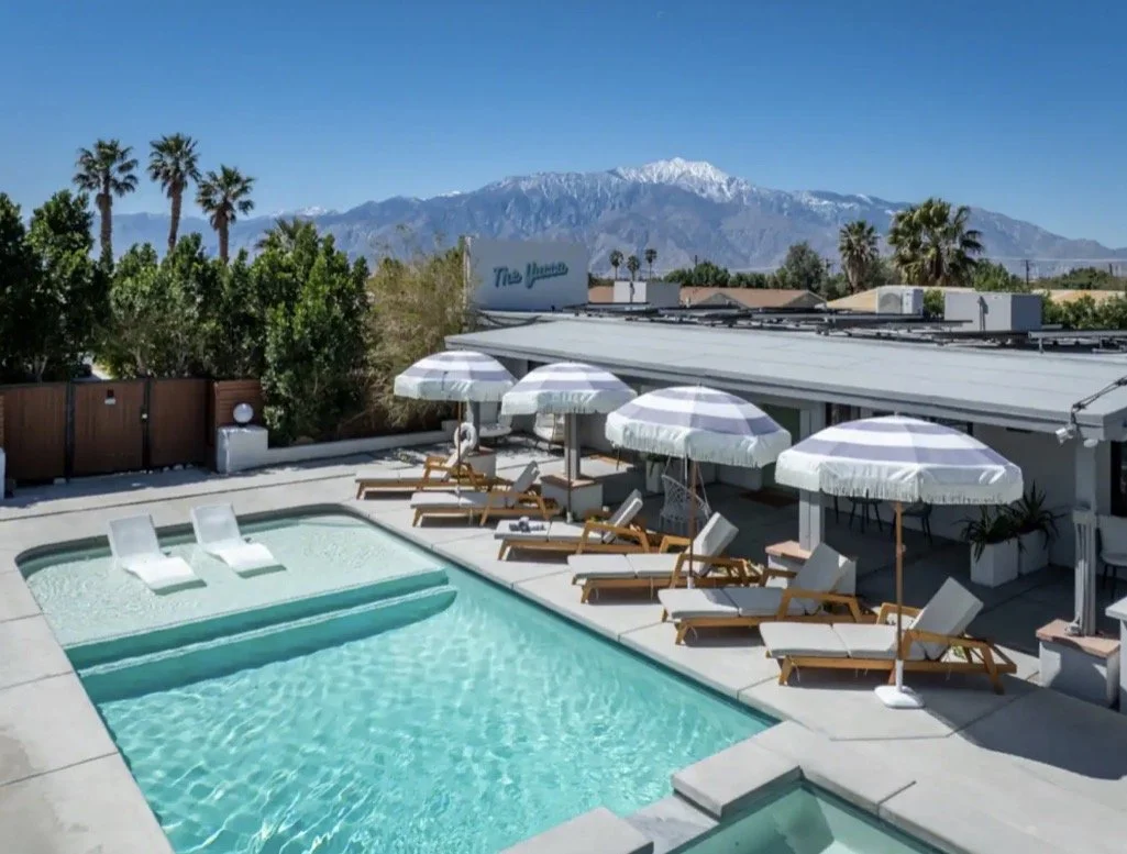 A rooftop pool area with lounge chairs and striped umbrellas, overlooking palm trees and snow-capped mountains in the background.