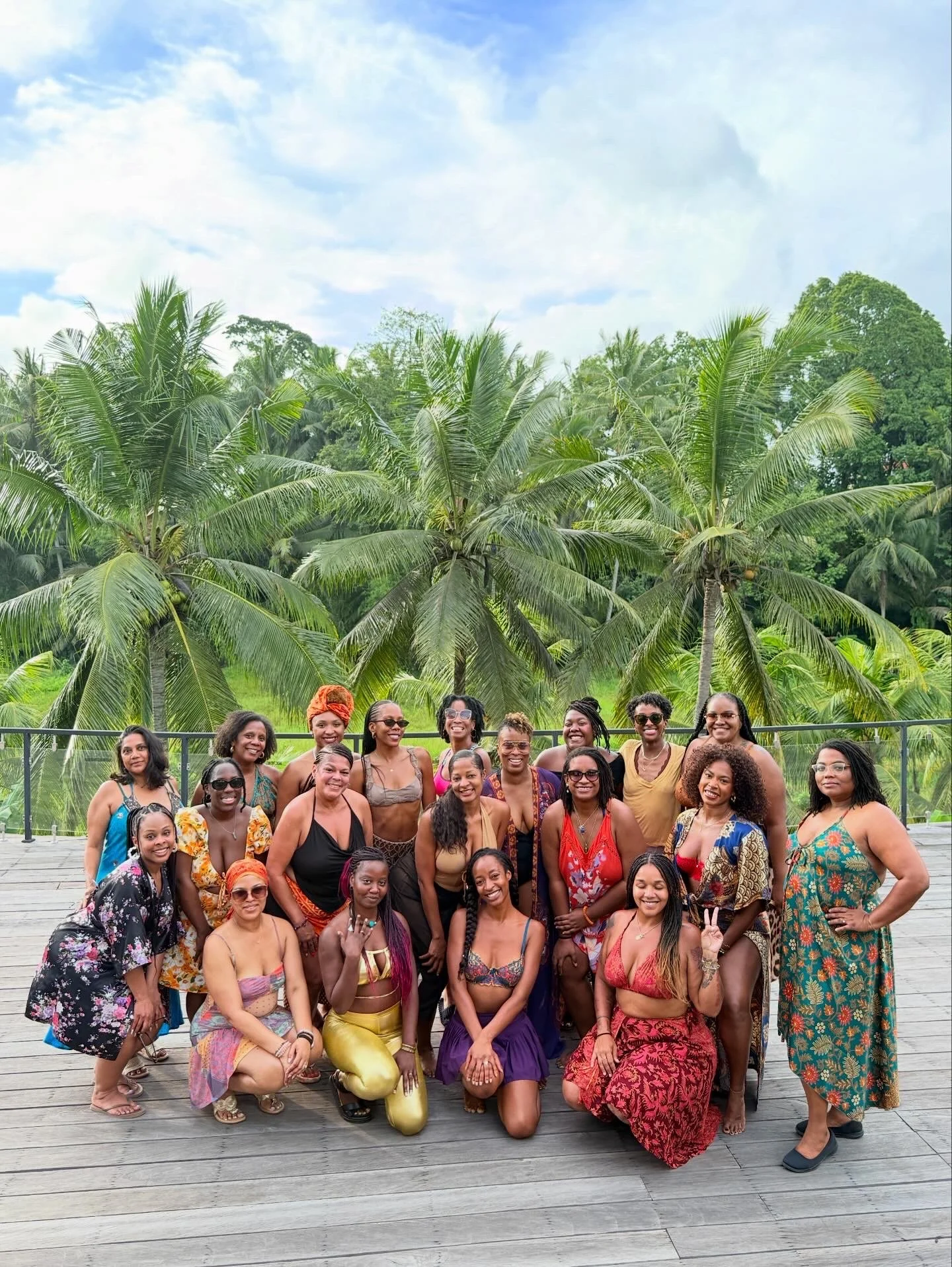 A group of women posing together outdoors on a wooden deck with lush green trees and palm trees in the background, under a partly cloudy sky.