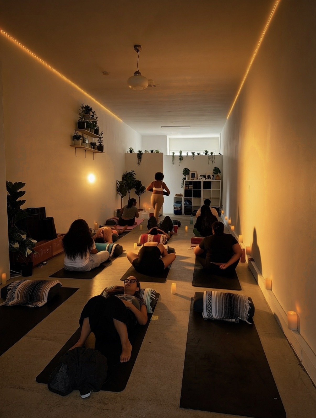A group of people participating in a yoga or meditation class in a cozy, dimly lit room with candles. The instructor is standing at the front, and the participants are lying or sitting on yoga mats.