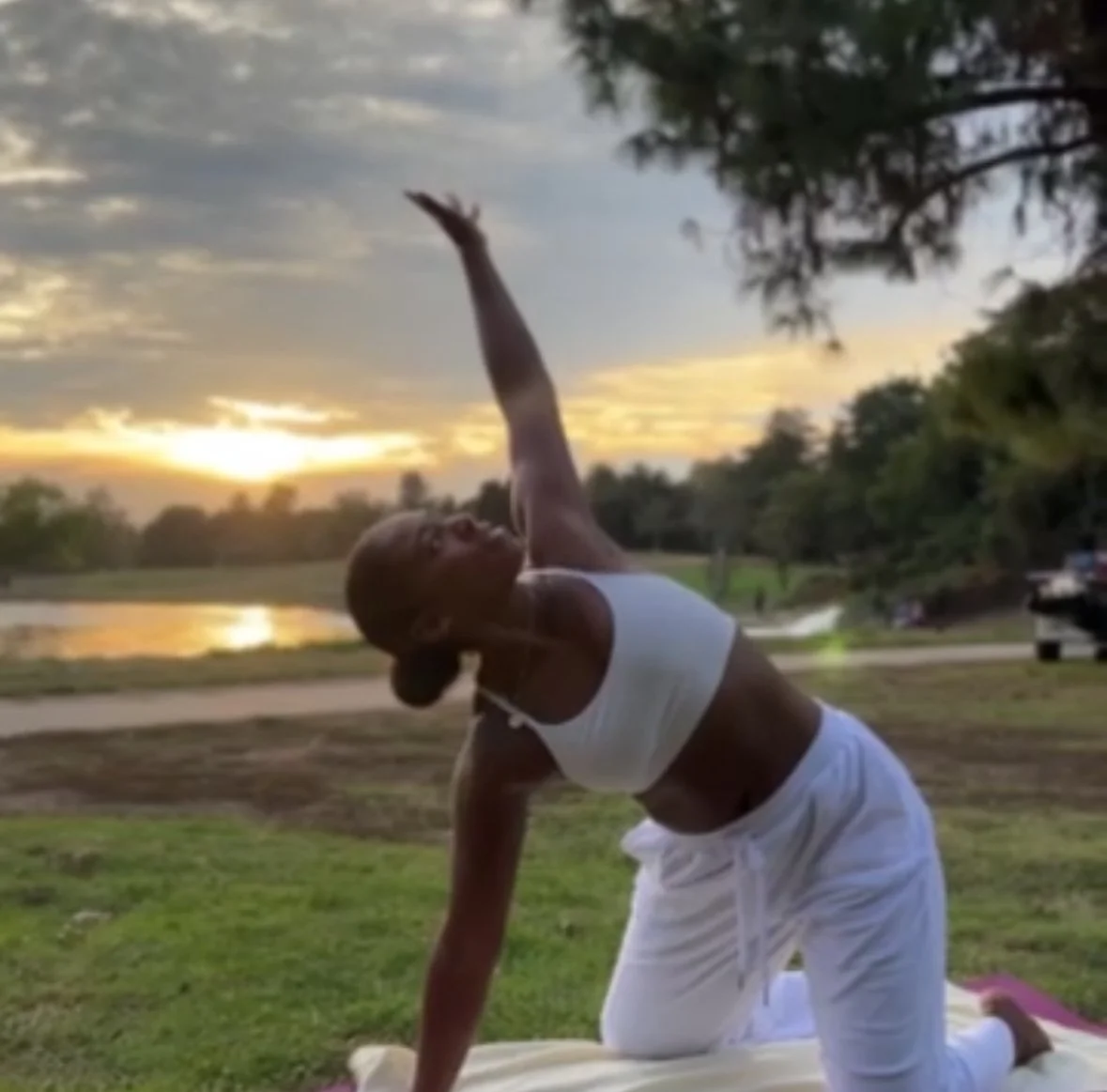 A woman practicing yoga outdoors on a mat during sunset, reaching upwards with her arm while kneeling on one knee amidst a grassy area with trees nearby.