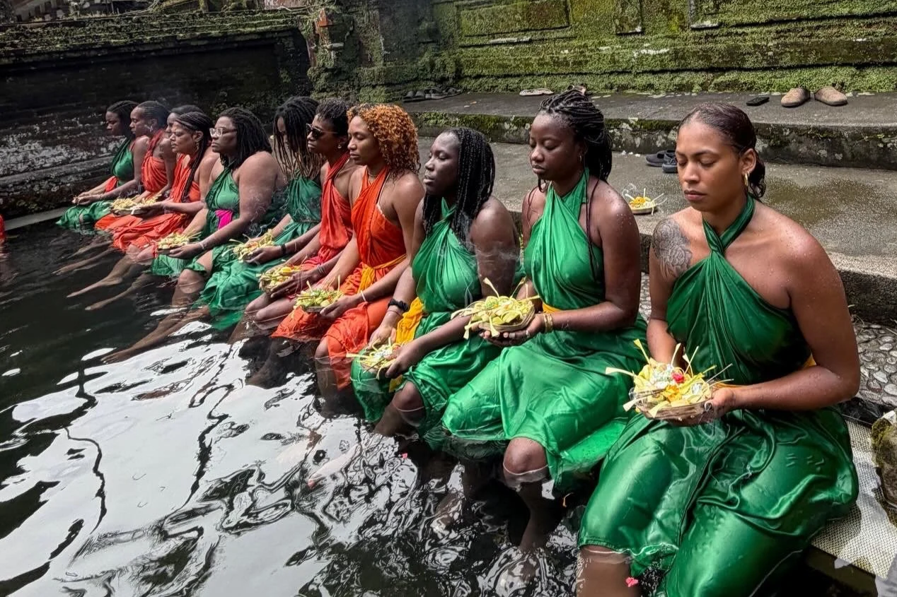 A group of women dressed in green and orange traditional attire sitting at the edge of a water body, holding offerings, during a cultural or religious ritual.