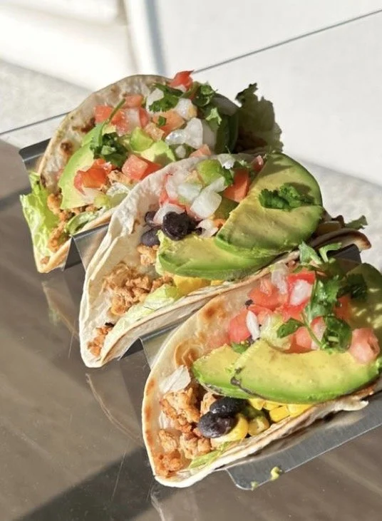 Three tacos filled with shredded meat, lettuce, diced tomatoes, avocado slices, and various vegetables, served on a metal tray.