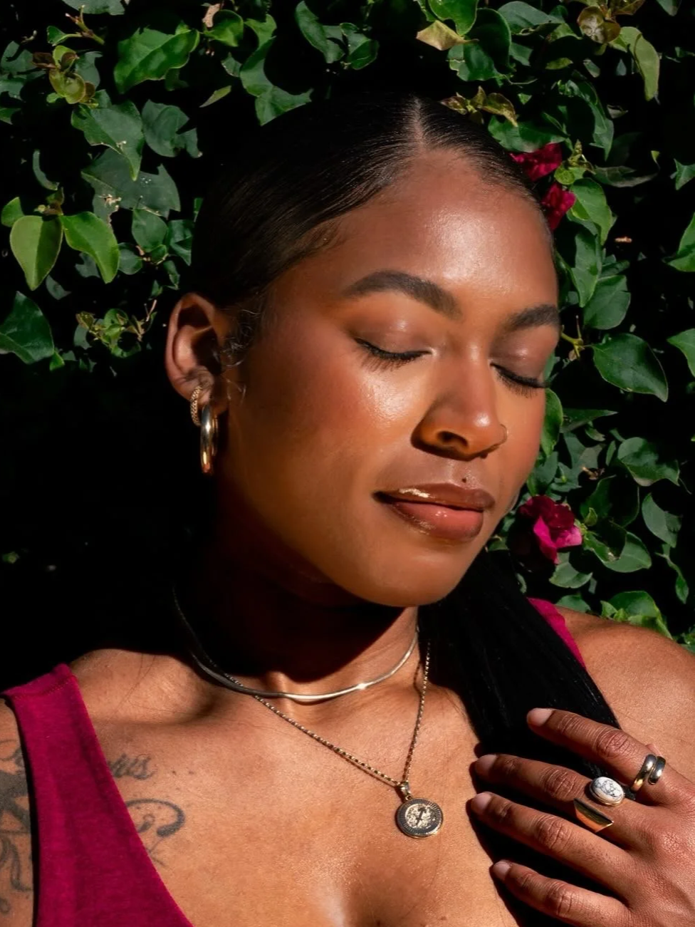 Close-up of a woman with closed eyes, wearing jewelry, and standing in front of lush green leaves and pink flowers.