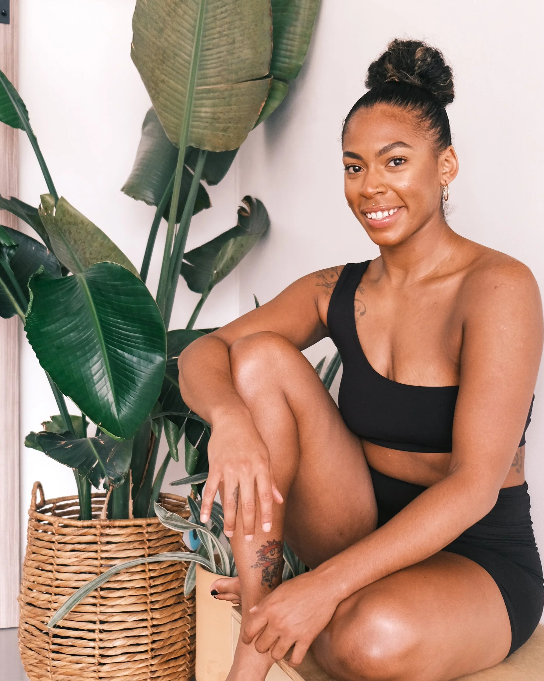 A woman with a top bun hairstyle, tattoos on her arm, earrings, and a nose ring, sitting beside a tall potted plant with large green leaves in a wicker basket, smiling at the camera.