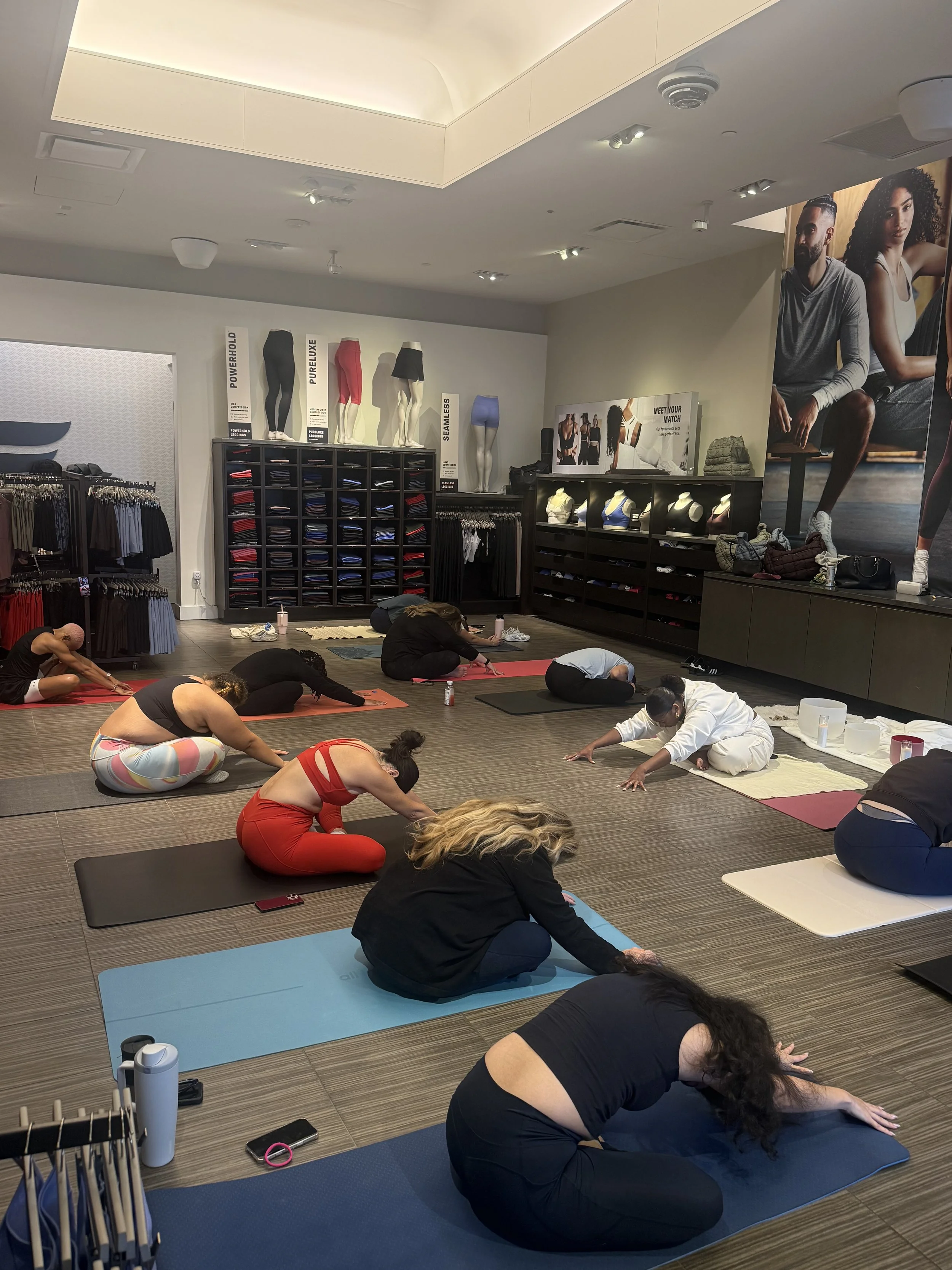 Group of people practicing yoga or stretching on mats in a fitness store or gym, with clothing displays and promotional posters on the wall.