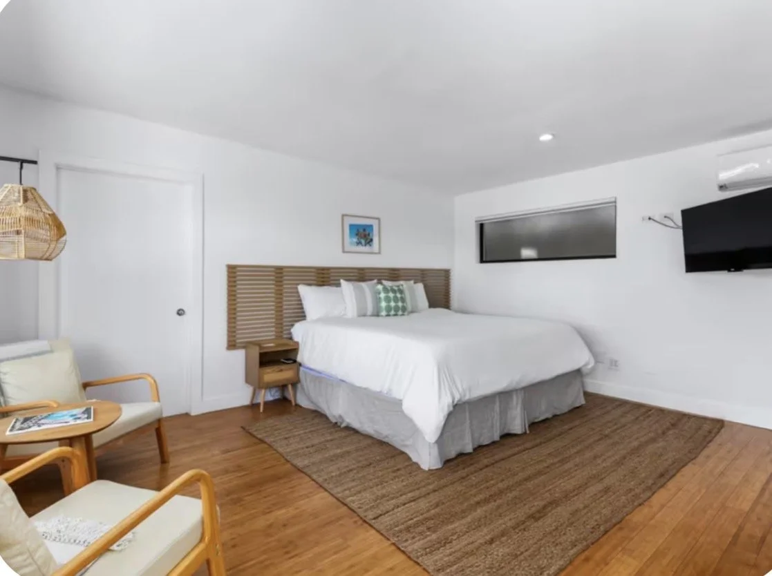 A hotel room with a made bed, a printed pillow, a wall-mounted TV, a window, and a sitting area with chairs and a table.
