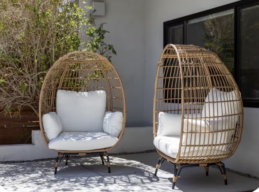 Two outdoor wicker egg chairs with white cushions on a patio, beside bushes and a window.