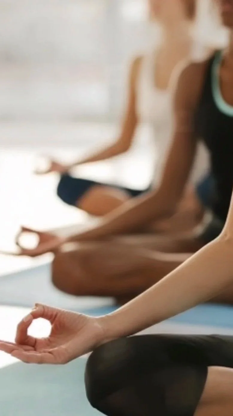 People practicing yoga in a studio, sitting on mats, in a meditative pose with hands in a mudra.
