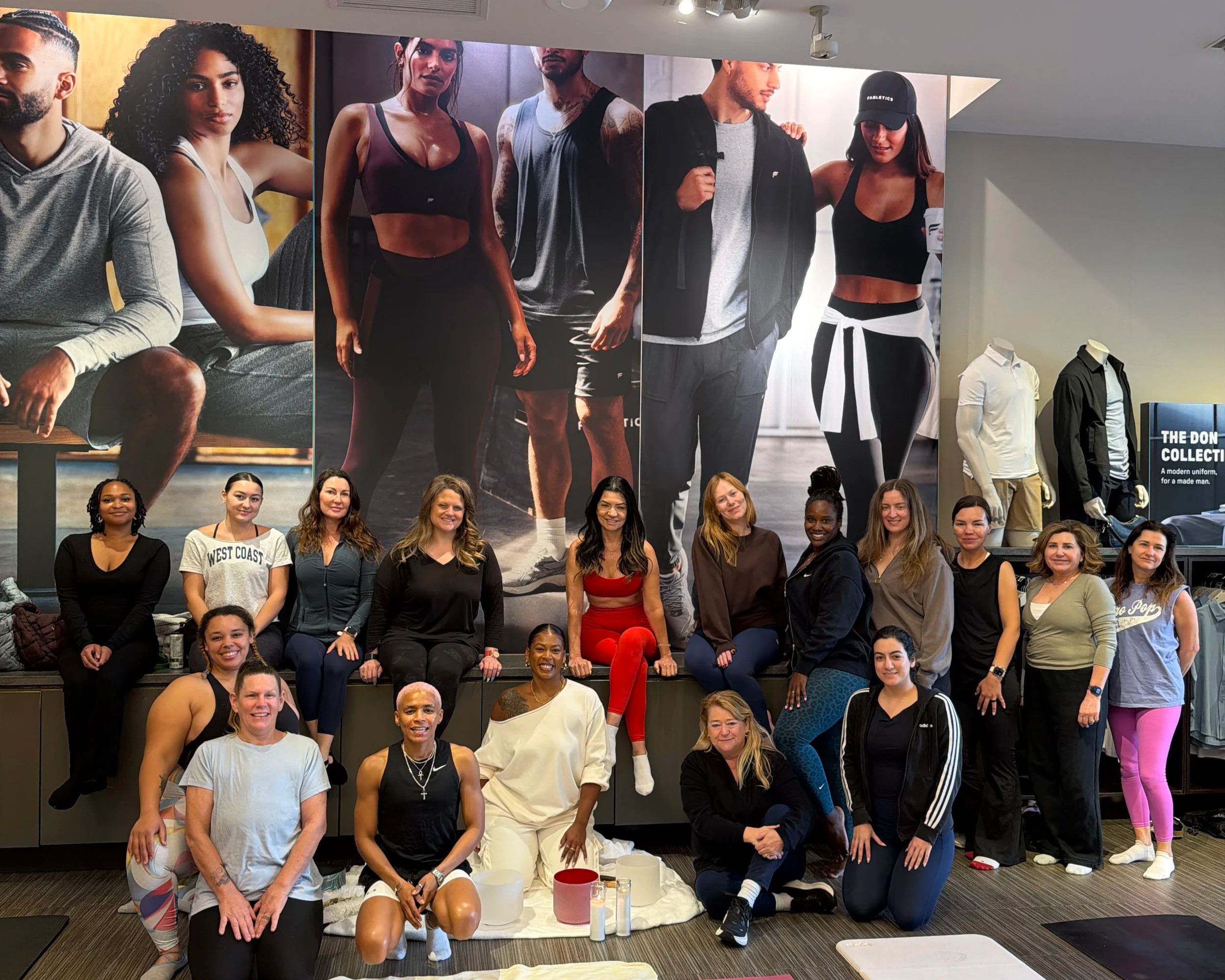 Large group of diverse women and men gathered in front of a wall with large fashion photographs, posing and smiling for a group photo in a casual indoor setting.