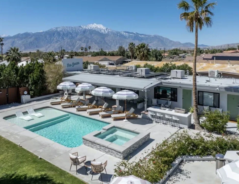 Pool area with lounge chairs and umbrellas, hot tub, surrounded by greenery and palm trees, with mountains in the background.