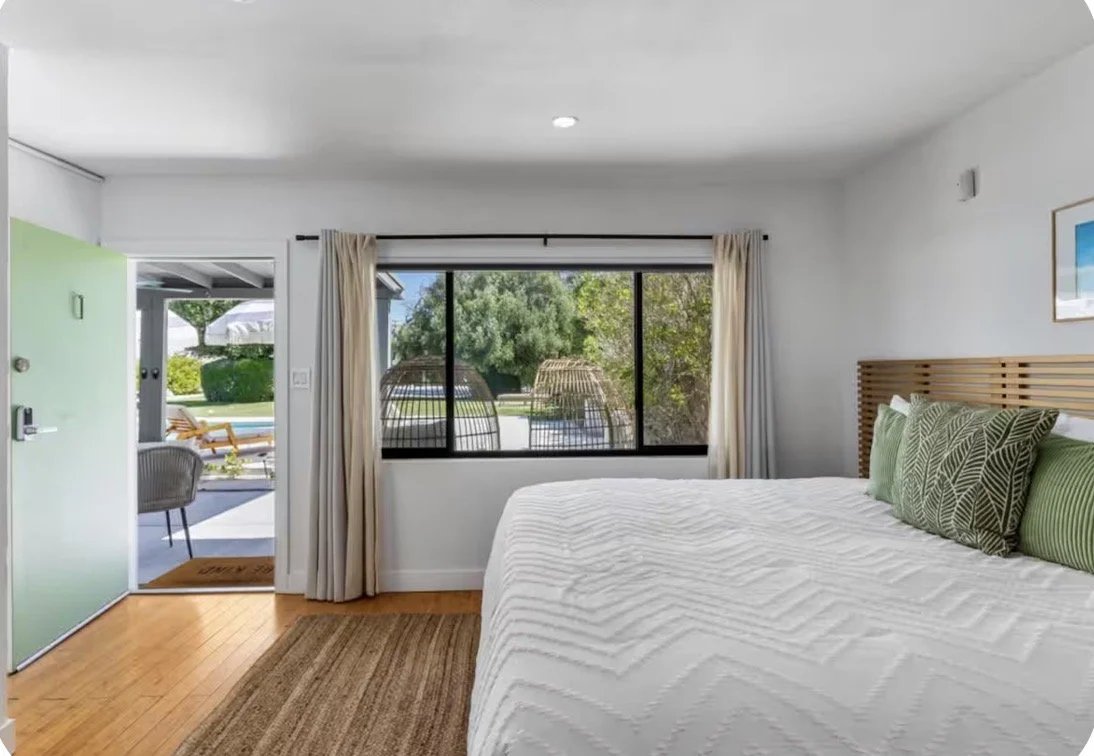 A bedroom with a large bed, green pillows, a wooden headboard, a window with a view of outdoor seating, and an open door leading outside to a patio with lounge chairs and greenery.
