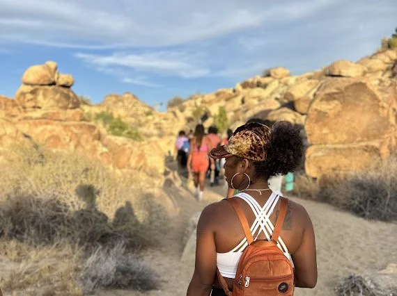 A woman with curly hair, wearing sunglasses and a backpack, hiking on a trail with other people in a desert landscape with rocky formations.
