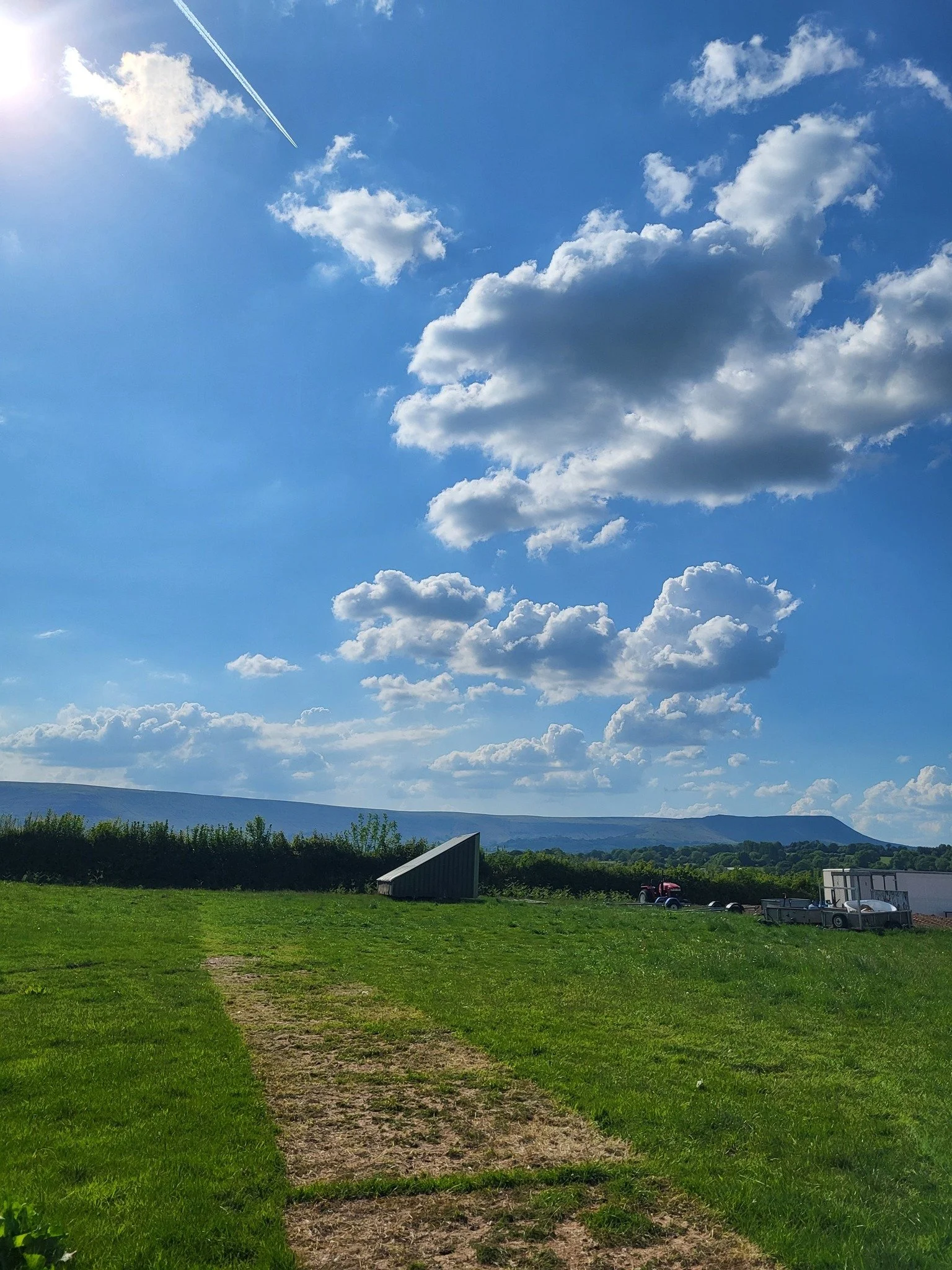A grassy field with a dirt path in the foreground, a blue sky with scattered clouds, and a mountain in the distance. There are trailers and equipment on the right side of the field.